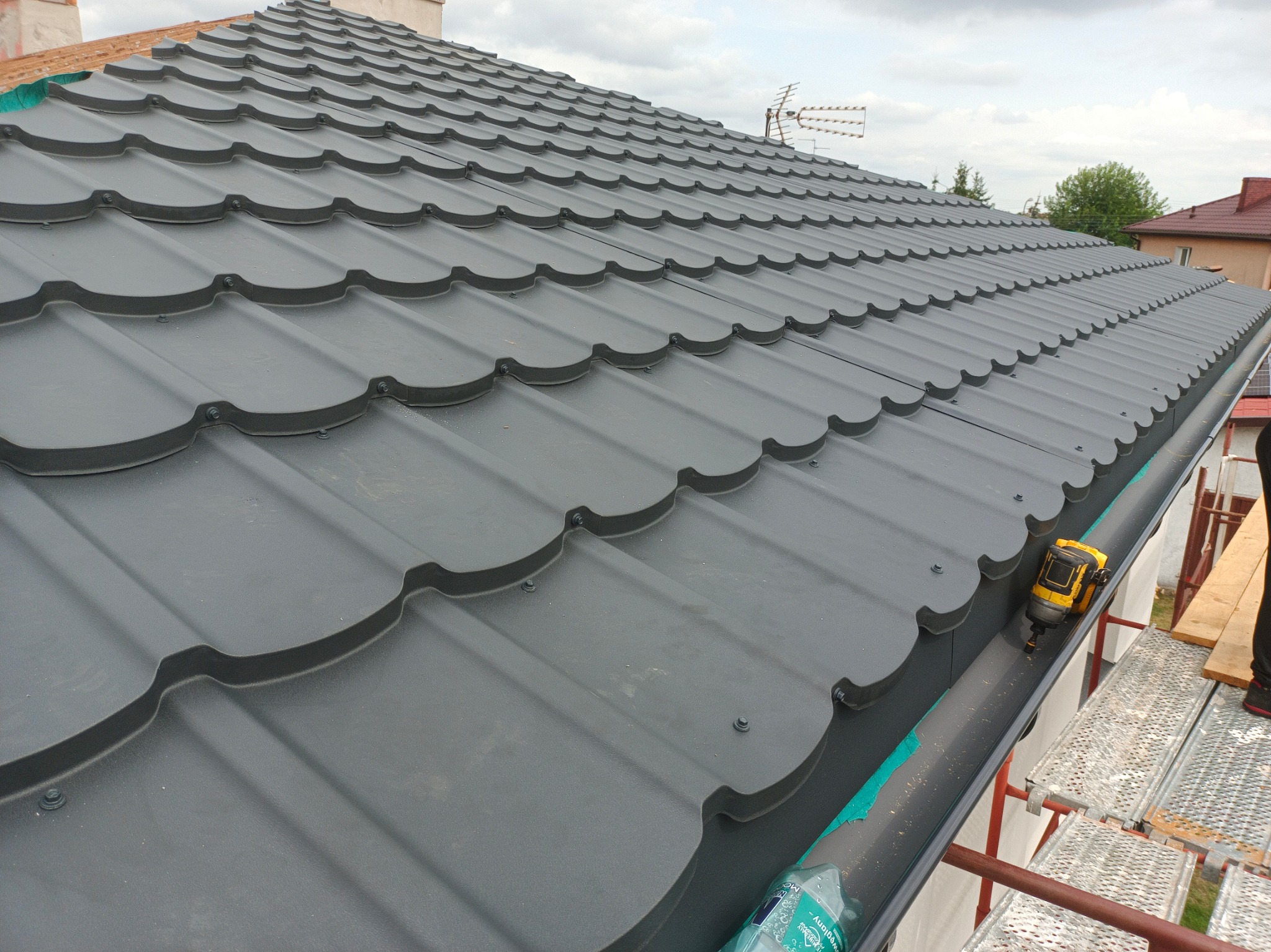 Nowy, grafitowy dach z blachodachówki modułowej, widok z rusztowania, z narzędziem do montażu rynien i butelką wody w tle.