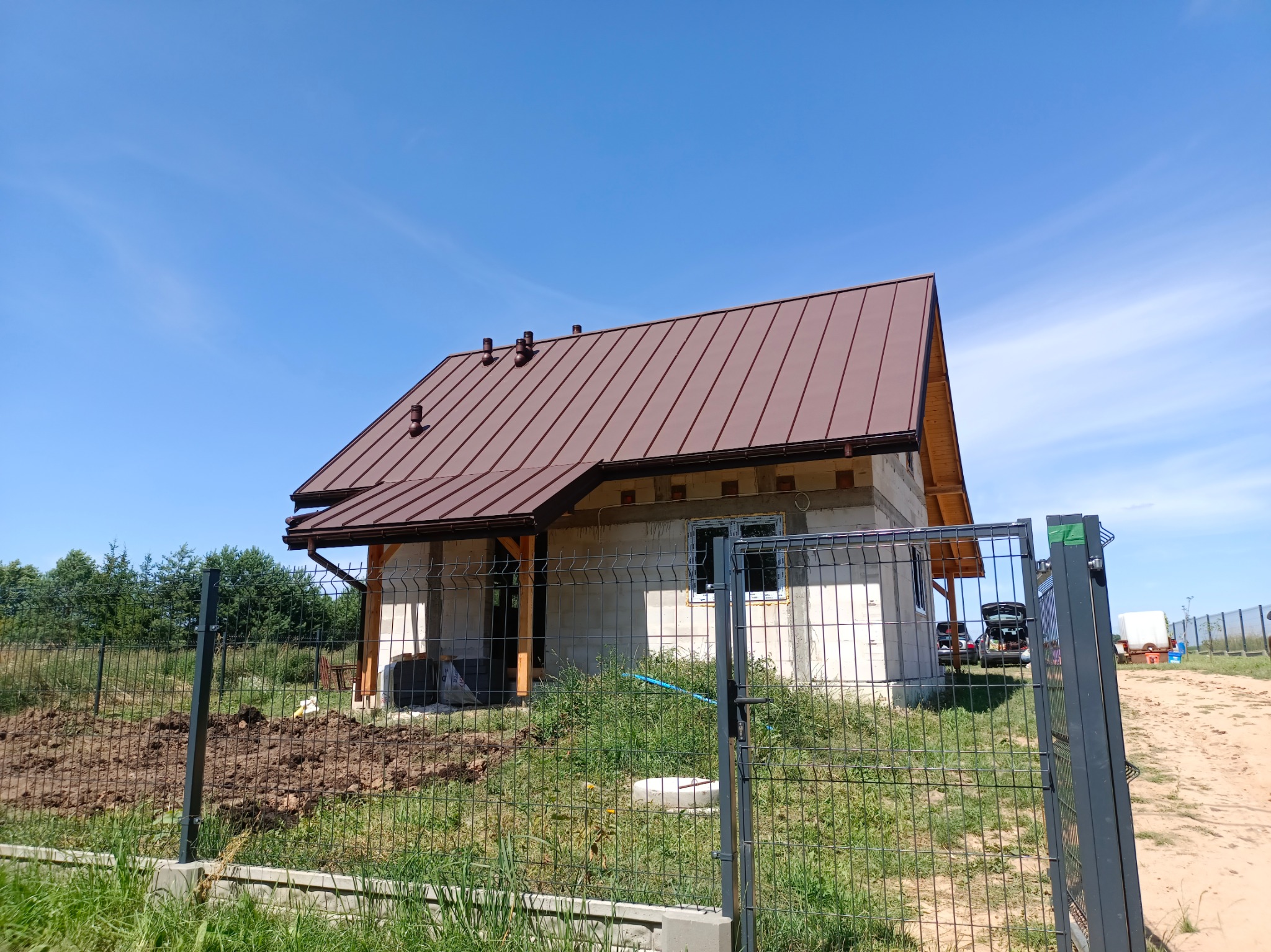 Niedokończony dom jednorodzinny z brązowym dachem z blachy na tle błękitnego nieba, widoczny przez metalowe ogrodzenie.