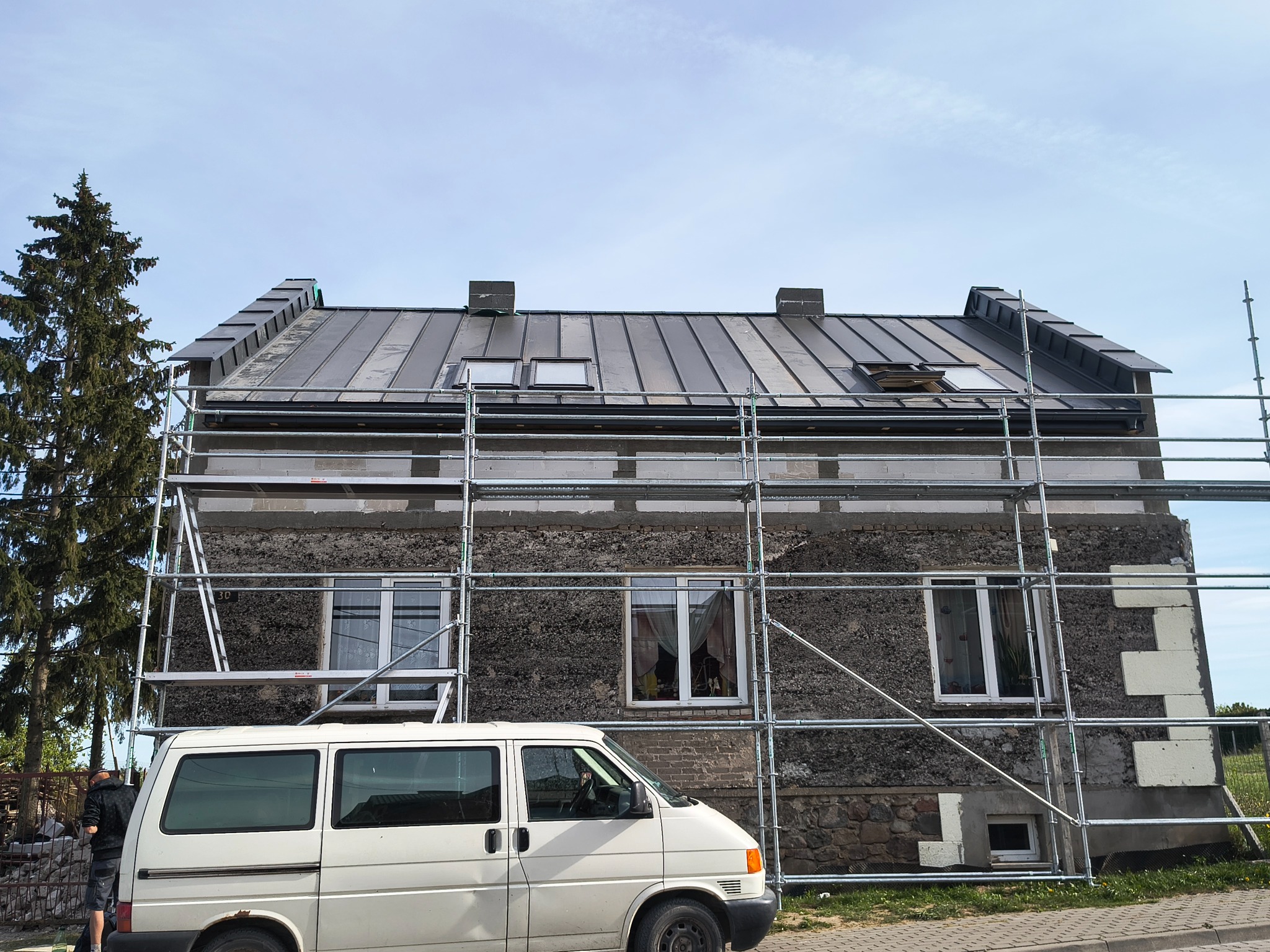 Dom w trakcie remontu dachu z blachy na rąbek stojący, widoczny z poziomu ulicy, z rusztowaniem i samochodem dostawczym. Częściowo ocieplona ściana szczytowa.