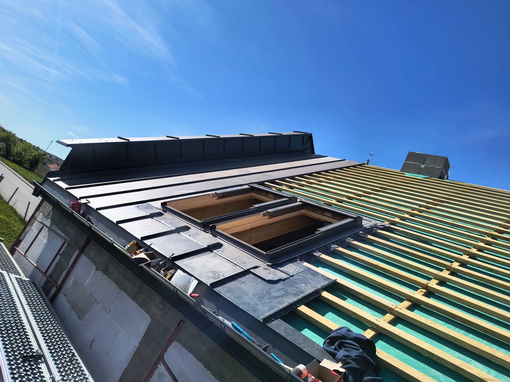 Montaż okien dachowych i blachy na dachu z widoczną konstrukcją drewnianą. Praca na wysokości, jasne niebo w tle. Częściowo pokryty dach z blachy.