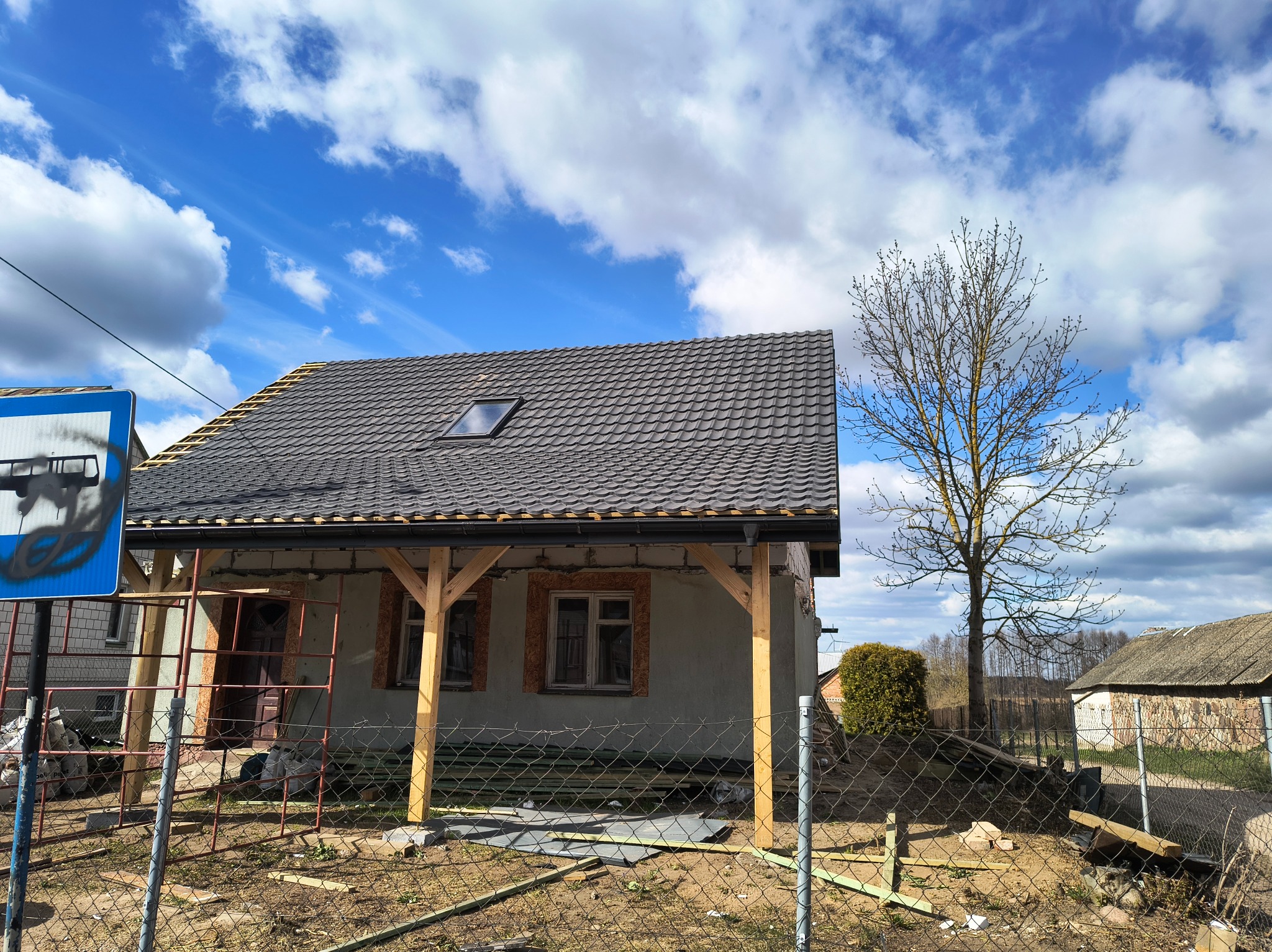 Dom z nowym, grafitowym dachem z dachówki modułowej i oknem połaciowym, w trakcie budowy werandy. W tle błękitne niebo z chmurami i drzewo bez liści.