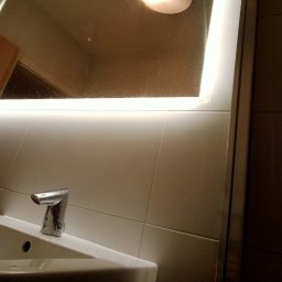 Perfekcyjna Pani Domu - Błyszcząca, chromowana bateria umywalkowa z automatycznym sensorem, zamontowana na białej umywalce w łazience z podświetlanym lustrem i beżowymi płytkami.