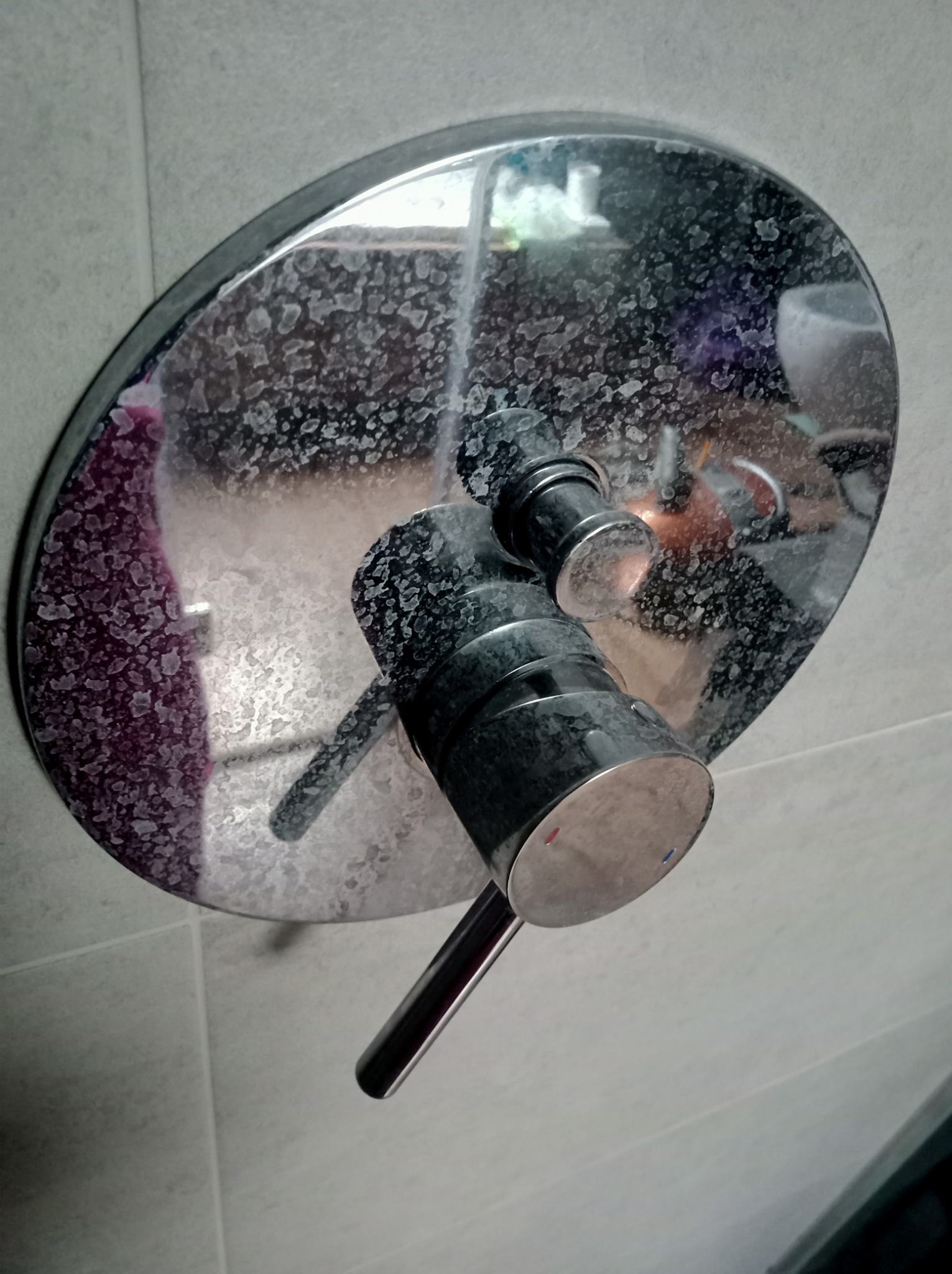 Zakamieniona, okrągła bateria prysznicowa z widocznymi osadami wapiennymi na chromowanej powierzchni i płytkach ceramicznych w tle.