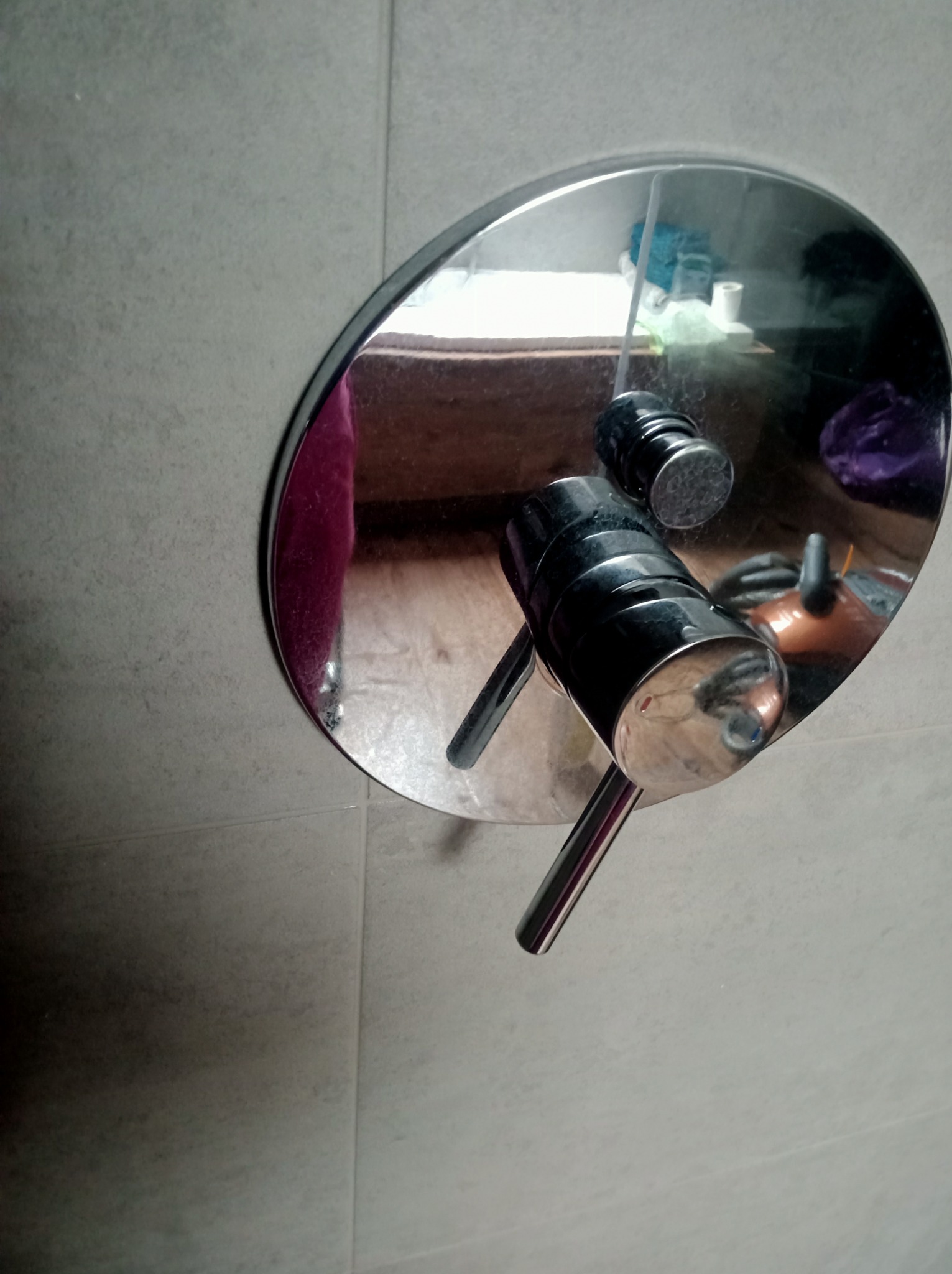 Bateria prysznicowa z okrągłą maskownicą, odbijająca fragment łazienki z ręcznikami i kosmetykami, zamontowana na szarej, kafelkowanej ścianie.