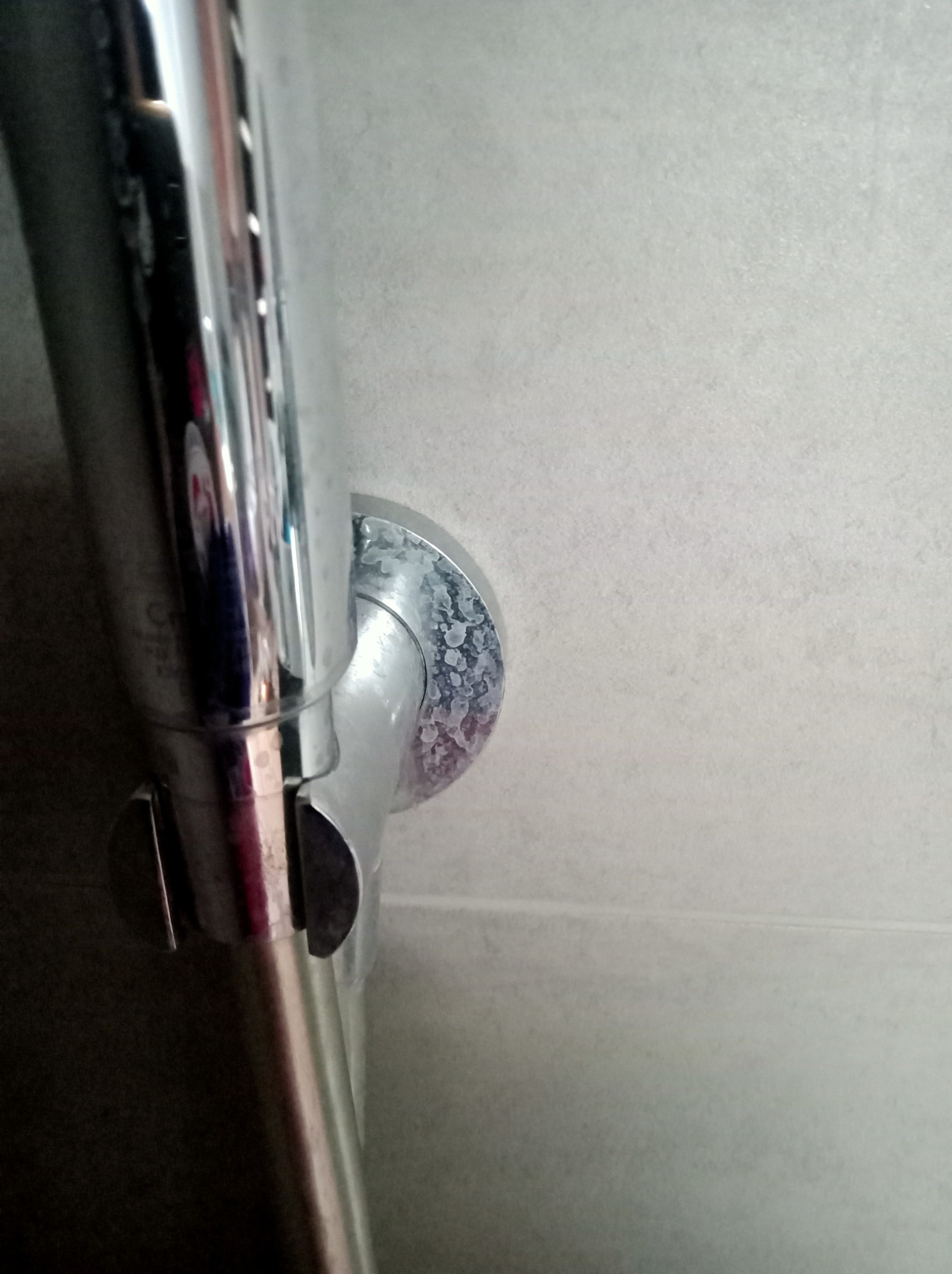 Chromowana rura prysznicowa z widocznym osadem kamiennym na podstawie mocującej do ściany, na tle jasnoszarej płytki.