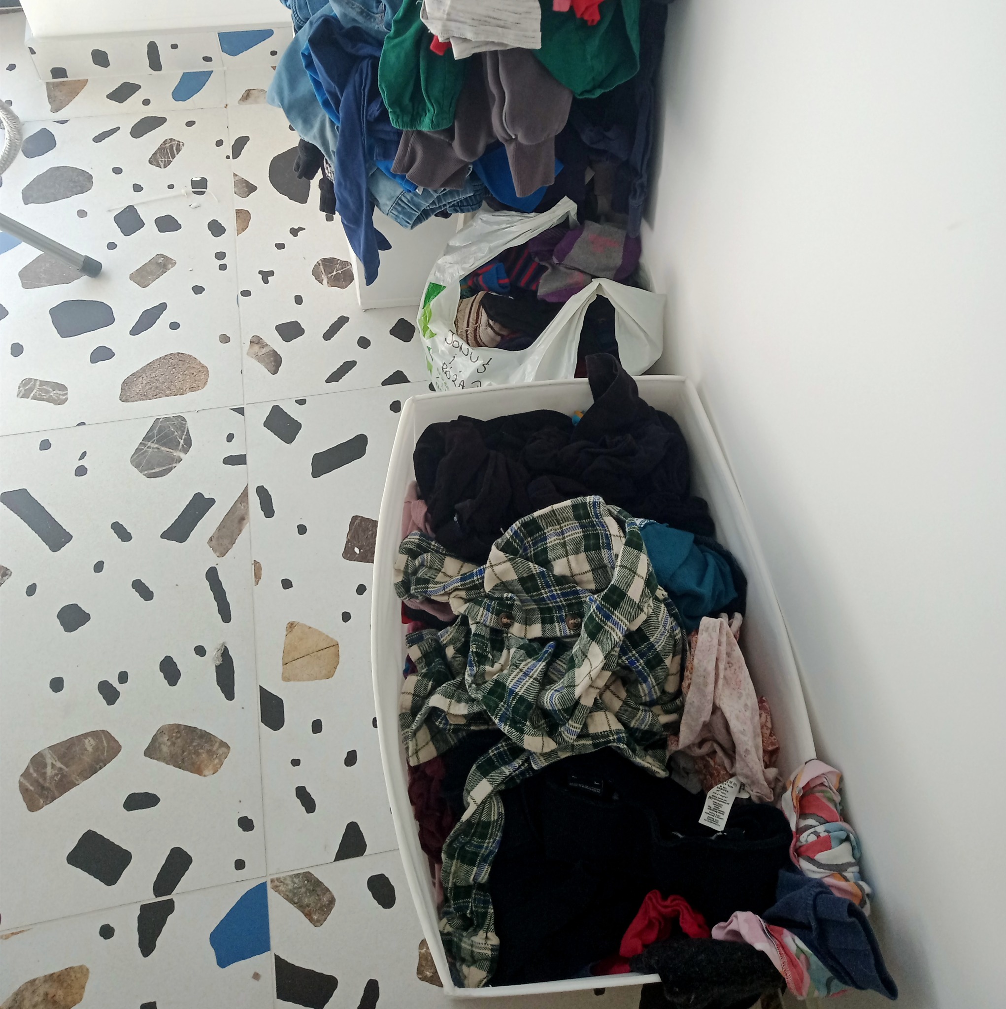 Dwa białe pojemniki i biała torba z ubraniami na podłodze z mozaiką w kolorach szarym, czarnym i beżowym, stojące przy białej ścianie.