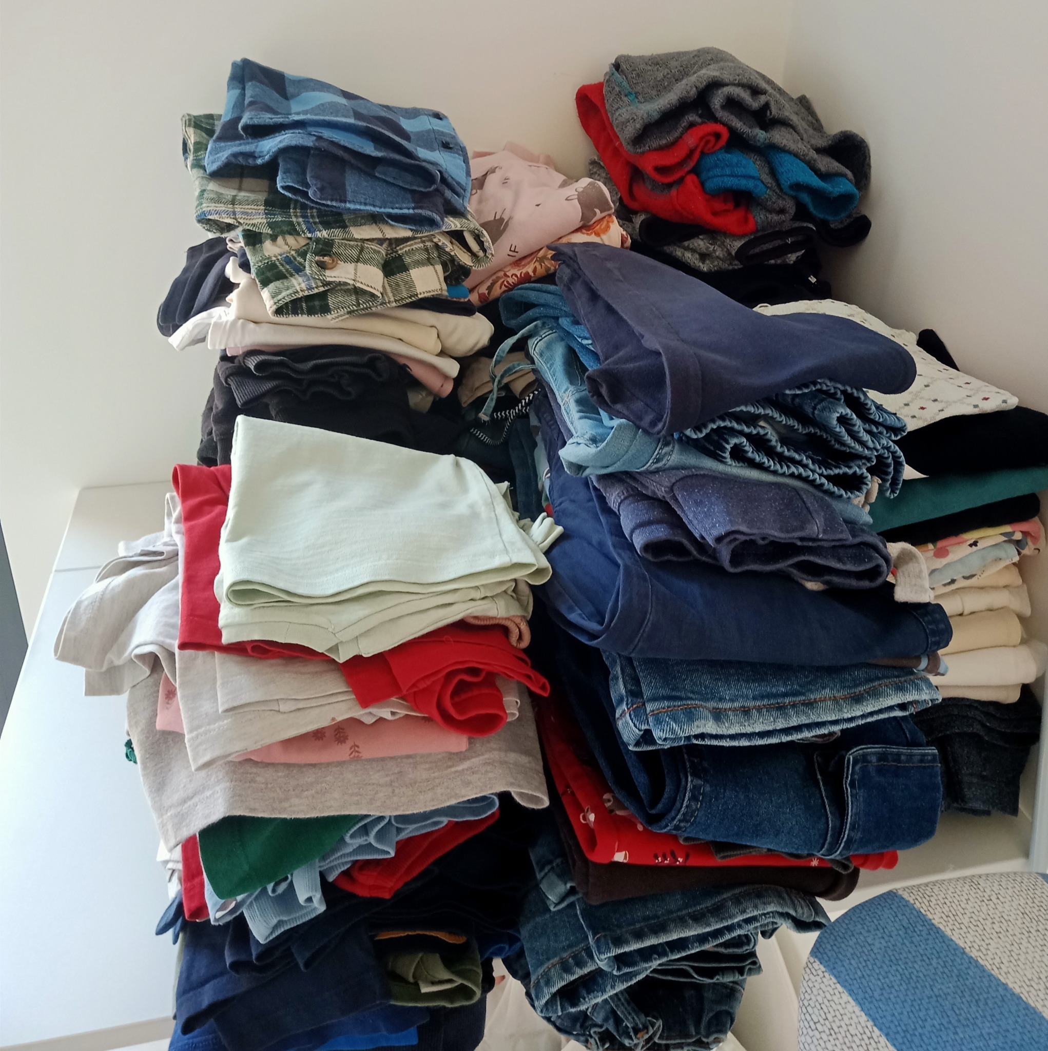 Stosy poskładanych ubrań w różnych kolorach i wzorach, w tym dżinsy, koszulki i flanelowe koszule, ułożone na białej półce.