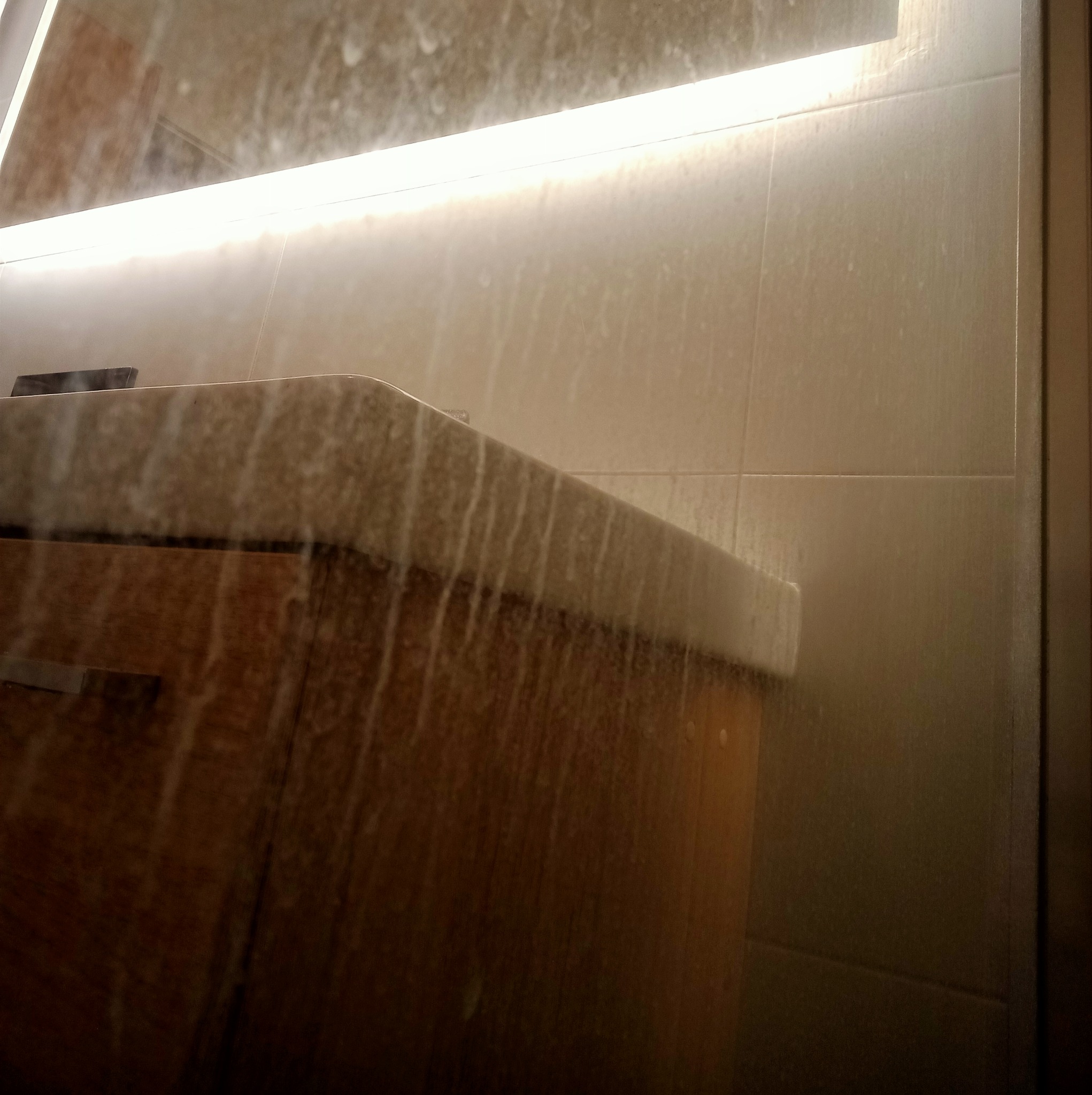 Zacieki po wodzie i mydle na umywalce z drewnianą szafką w łazience z podświetlanymi płytkami.