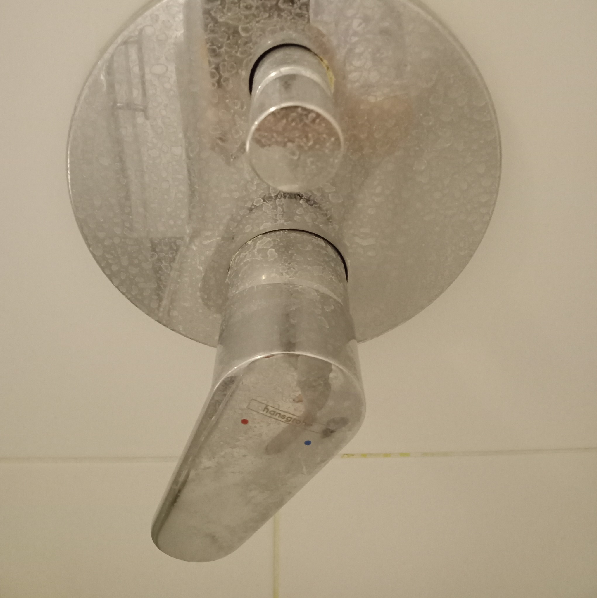 Chromowana bateria prysznicowa Hansgrohe pokryta osadem z kamienia na białej płytce.