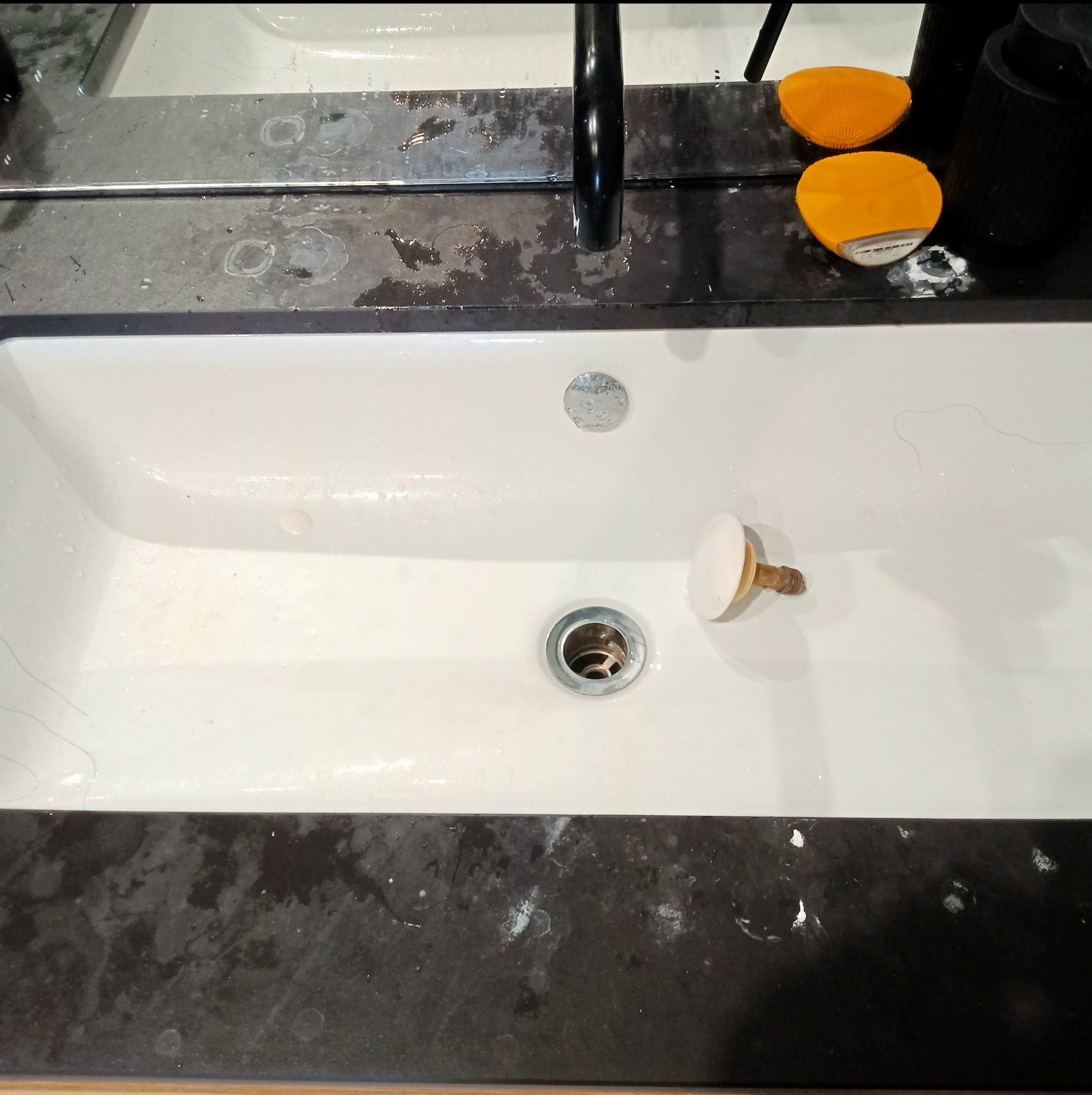 Biała umywalka z chromowanym odpływem, brudnym korkiem i śladami po wodzie na czarnym blacie oraz odbiciem w lustrze