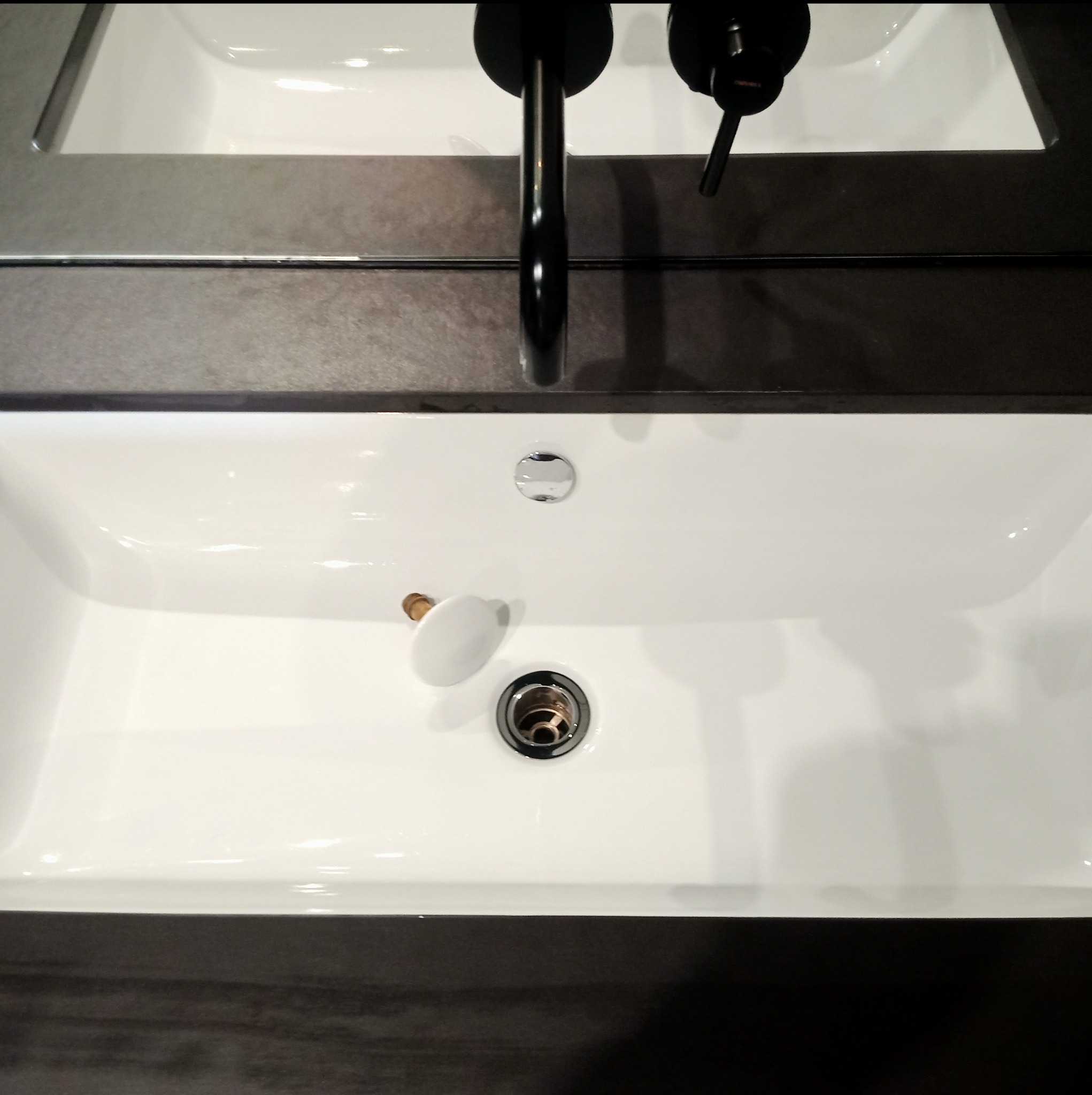 Biała, prostokątna umywalka z czarną baterią i pop-up korkiem w łazience z ciemnoszarym blatem.