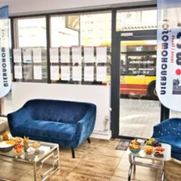 Wnętrze biura nieruchomości z niebieskimi sofami, stolikami kawowymi z przekąskami i flagami reklamowymi firmy West Nieruchomości, widok na ulicę z autobusem za szybą.