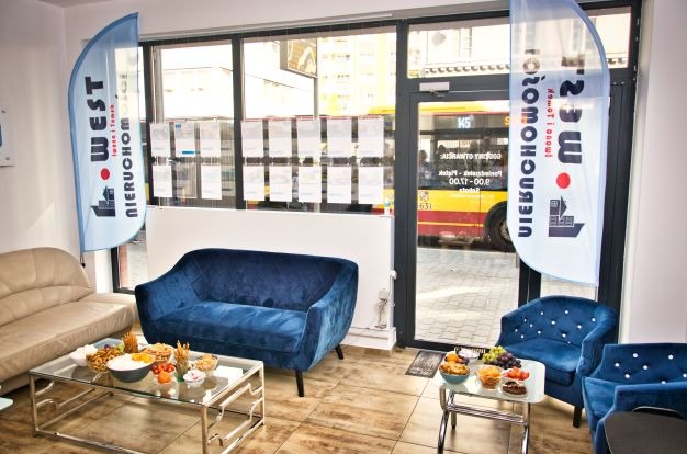 Wnętrze biura nieruchomości z niebieskimi sofami, stolikami kawowymi z przekąskami i flagami reklamowymi firmy West Nieruchomości, widok na ulicę z autobusem za szybą.