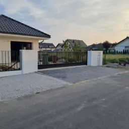 Nowoczesna posesja z przesuwną bramą wjazdową z czarnych, metalowych profili, osadzoną między białymi słupkami, z kostką brukową przed wjazdem i szarą nawierzchnią z kamyczków po lewej stronie.