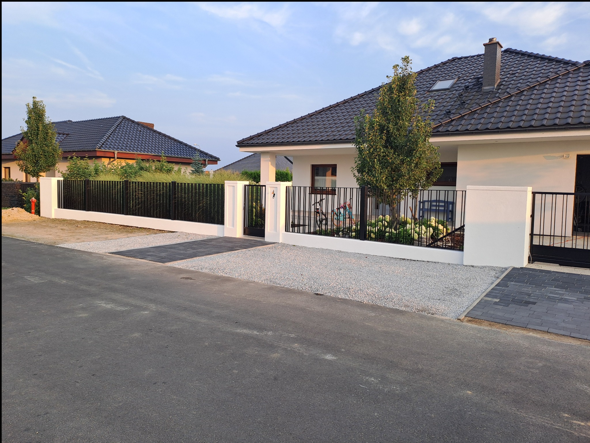 Nowoczesne ogrodzenie metalowe z bramą wjazdową, białe słupki, czarne przęsła, żwirowe podłoże, dom z ciemną dachówką w tle.
