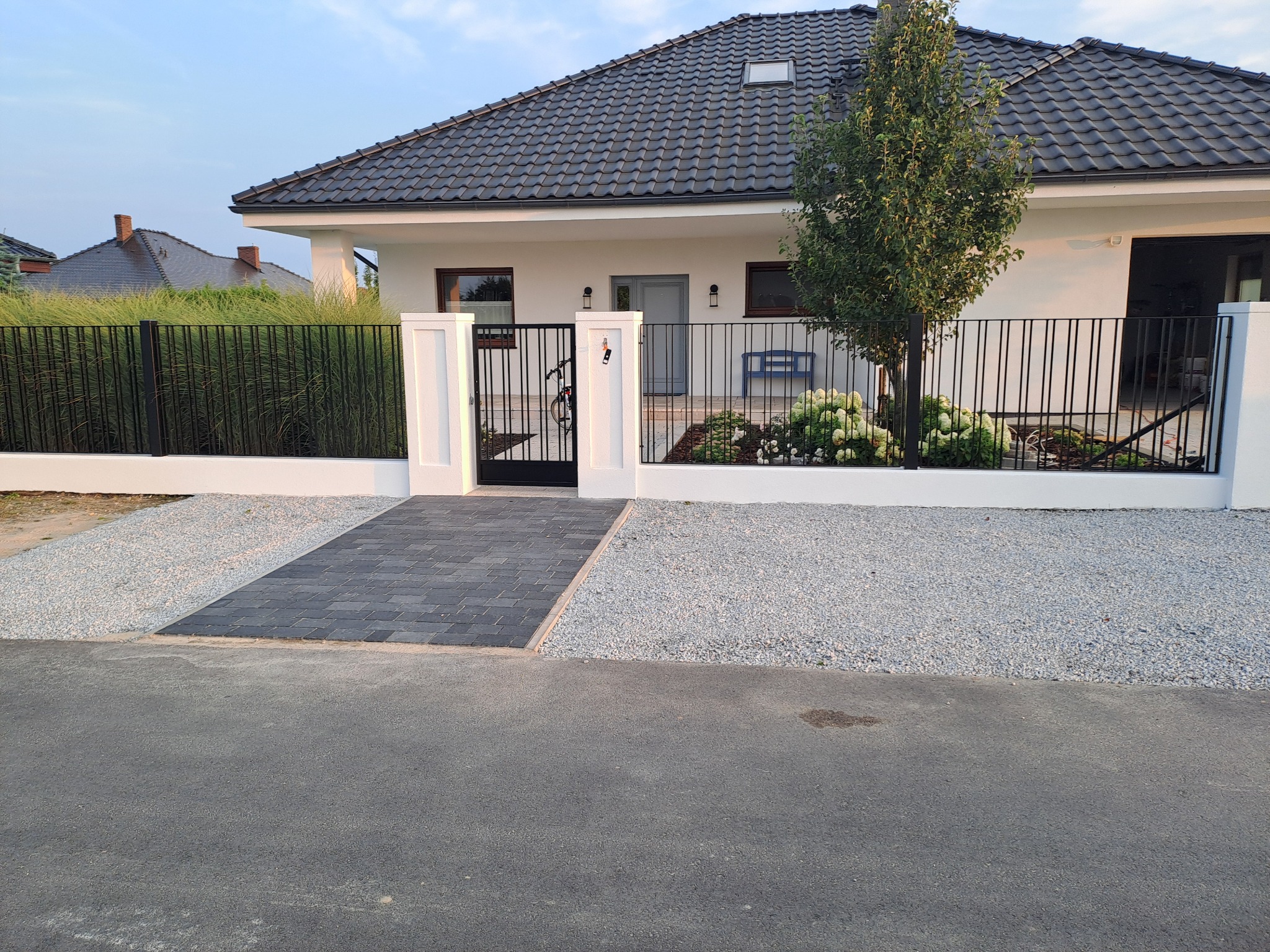 Nowoczesny dom jednorodzinny z czarnym ogrodzeniem sztachetowym na niskim murku, szara kostka brukowa i jasny żwir na podwórku, brama wejściowa z rowerem opartym o słupek.