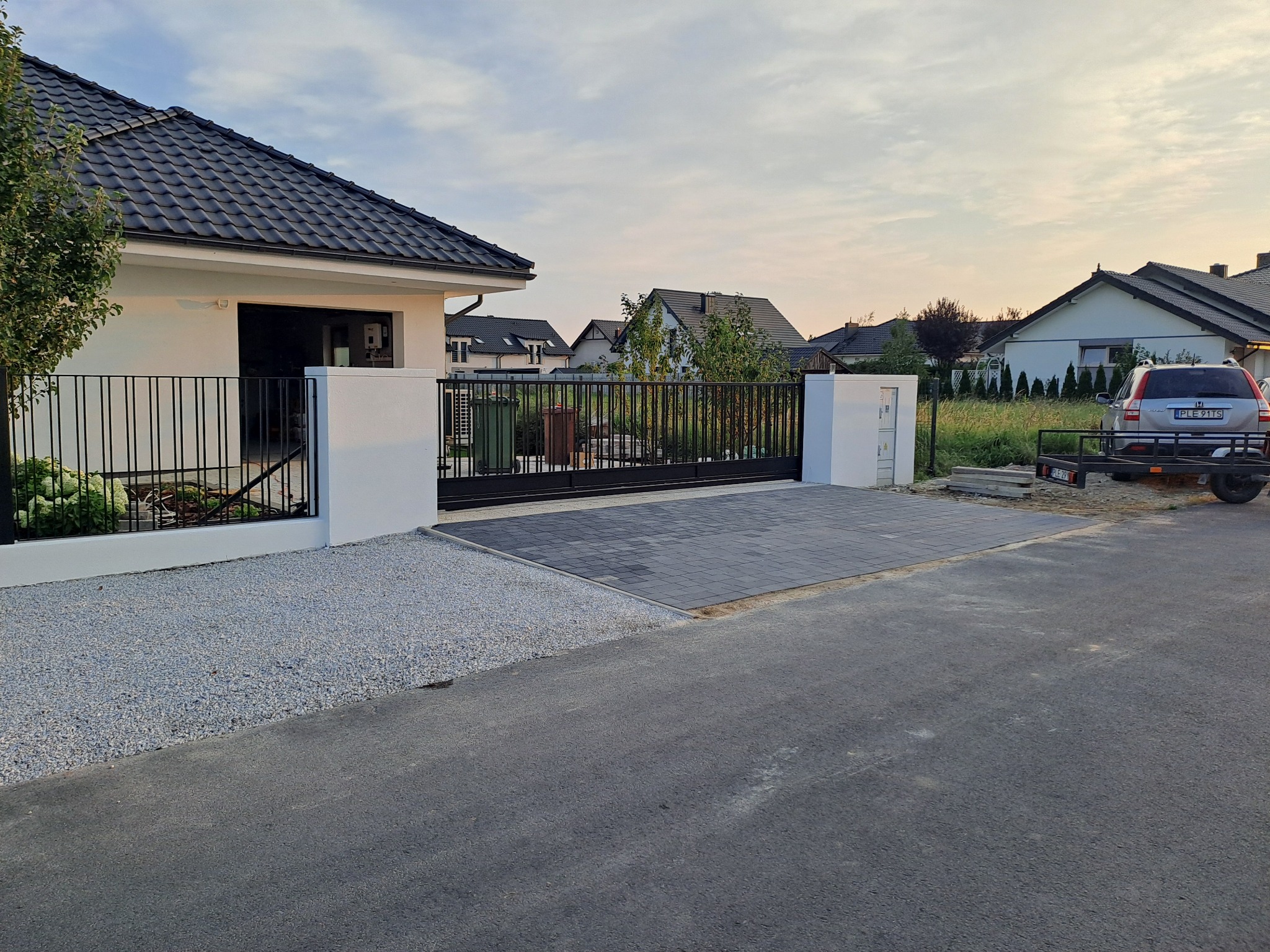 Nowoczesna posesja z przesuwną bramą wjazdową z czarnych, metalowych profili, osadzoną między białymi słupkami, z kostką brukową przed wjazdem i szarą nawierzchnią z kamyczków po lewej stronie.
