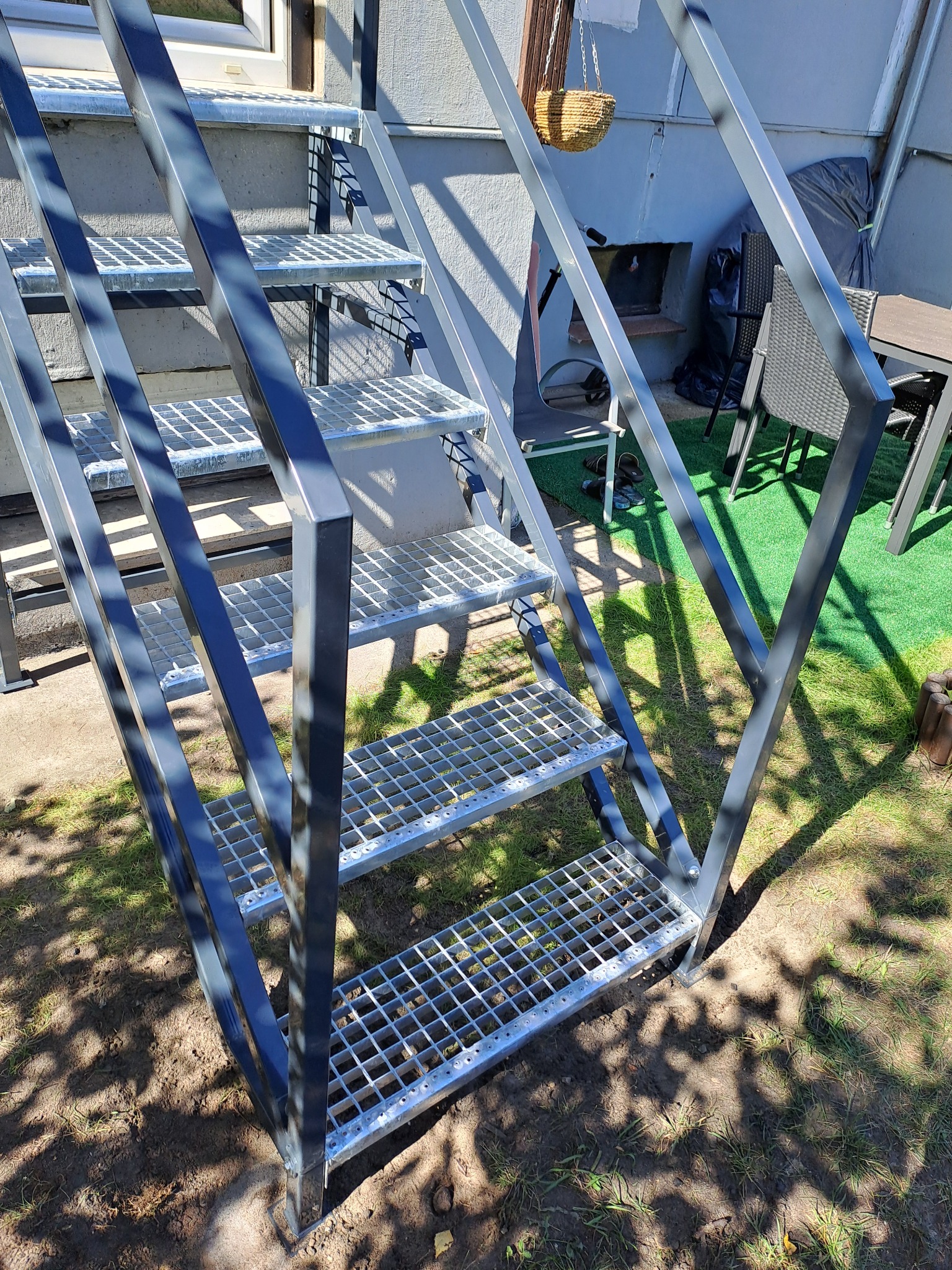 Zewnętrzne metalowe schody z kratowymi stopniami, prowadzące do wejścia na podwyższeniu, widoczne z dołu na tle trawnika i ściany budynku.