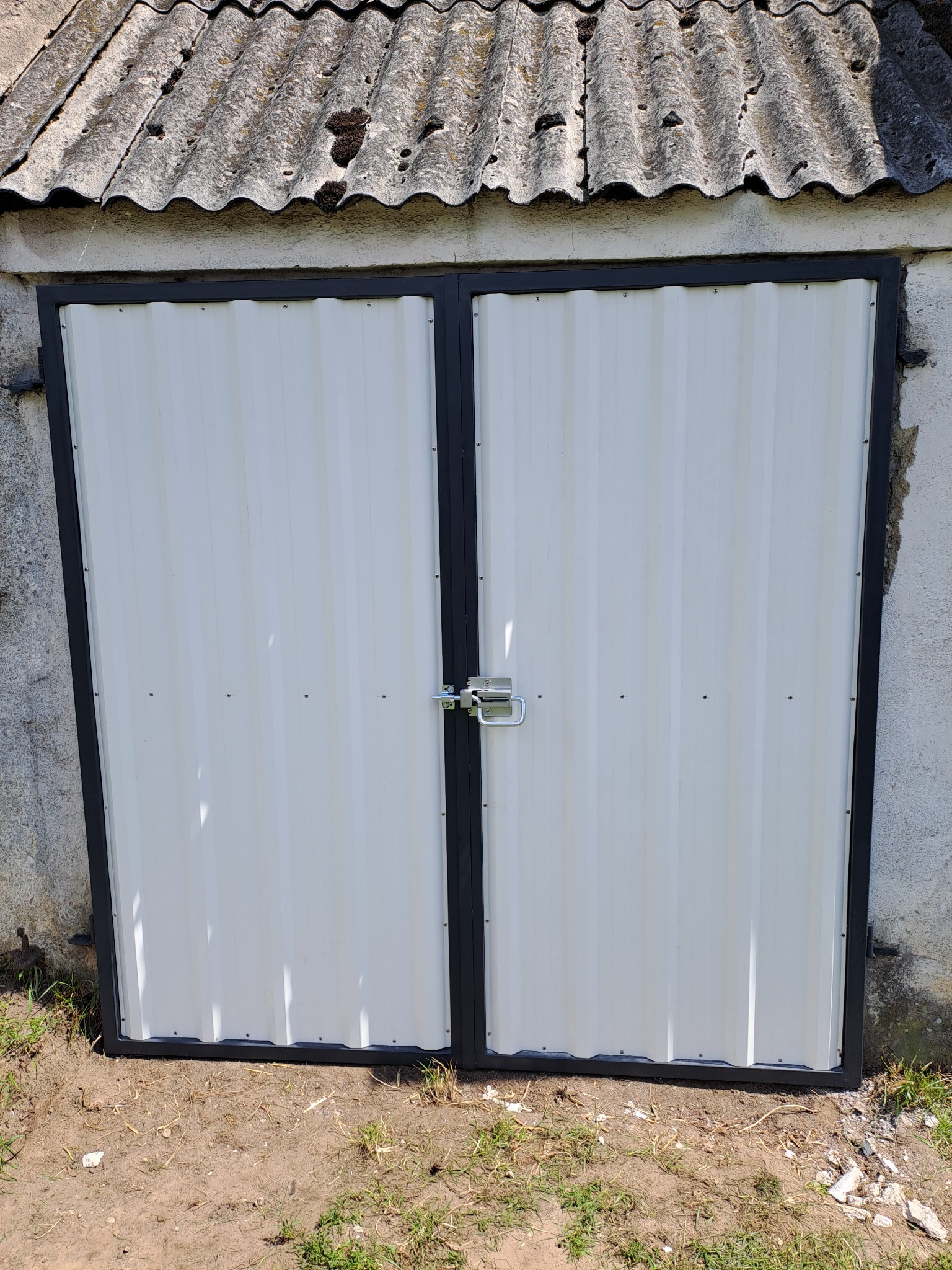 Dwuskrzydłowa brama z blachy trapezowej, pomalowana na biało z czarną ramą, zabezpieczona kłódką, widoczna na tle starego budynku gospodarczego w Bojanicach.