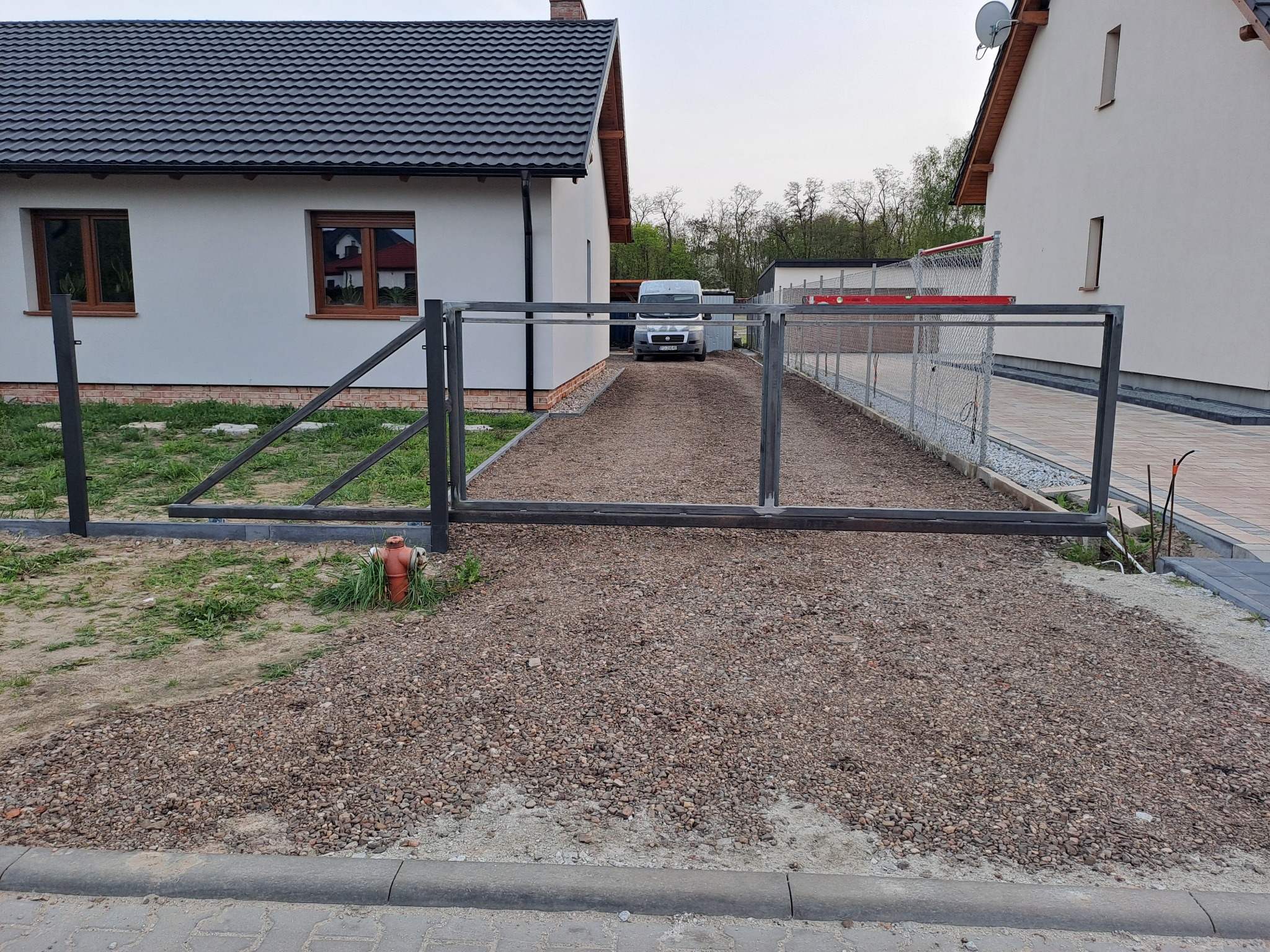 Surowa metalowa konstrukcja bramy wjazdowej z widocznym fragmentem ogrodzenia, przed budynkiem z szarą elewacją i brązowymi oknami, na żwirowym podłożu, z samochodem dostawczym w tle.