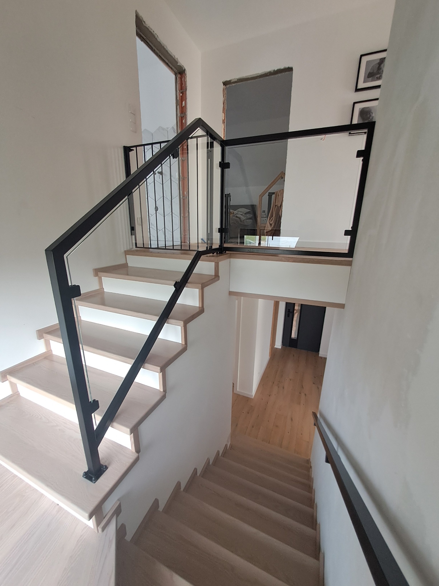 Nowoczesna balustrada ze szkła i czarnego metalu zamontowana przy drewnianych schodach w jasnym wnętrzu domu, widok z góry schodów.