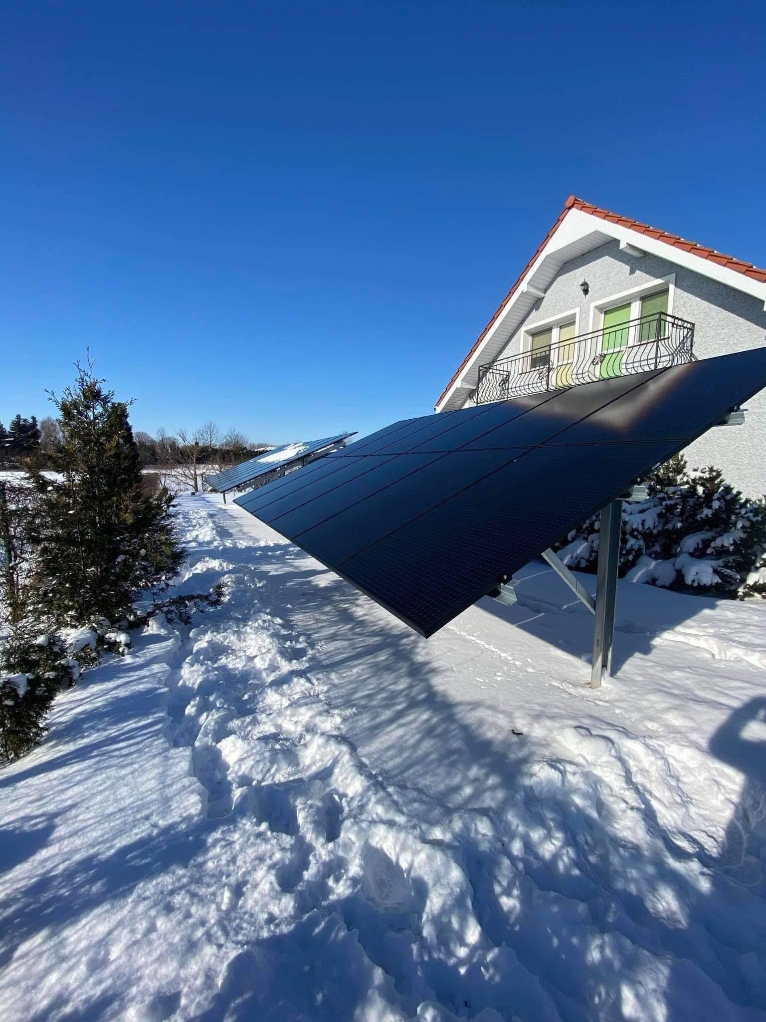 Rząd paneli słonecznych pokrytych śniegiem w zimowym krajobrazie, z domem z balkonem w tle i śladami stóp w śniegu prowadzącymi wzdłuż instalacji.