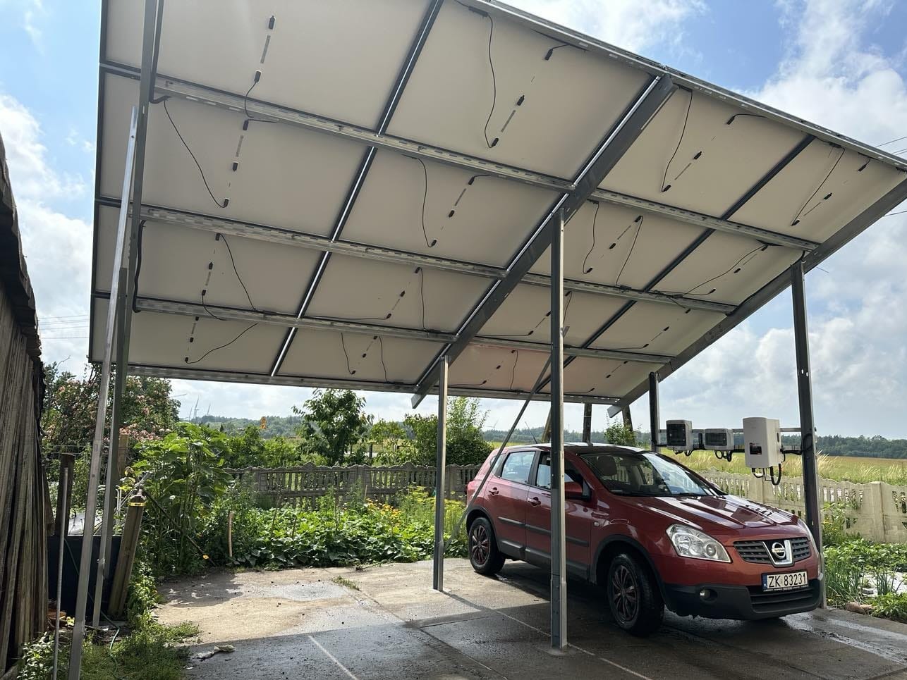 Czerwony samochód Nissan Qashqai zaparkowany pod zadaszeniem z paneli fotowoltaicznych, na tle wiejskiego krajobrazu z polami i zielenią.