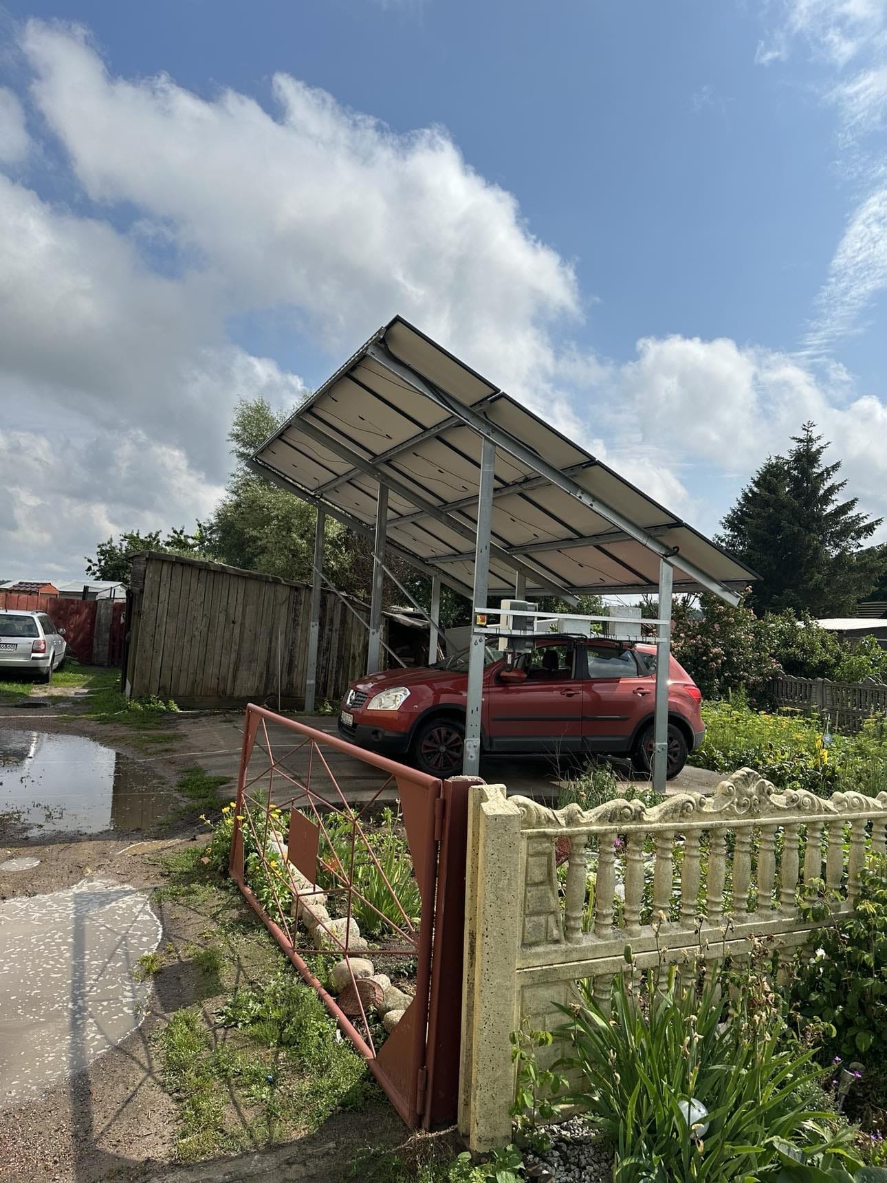 Czerwony samochód zaparkowany pod wiatą z paneli słonecznych w wiejskim otoczeniu, widoczne kałuże, otwarte metalowe ogrodzenie i zaniedbany ogród.
