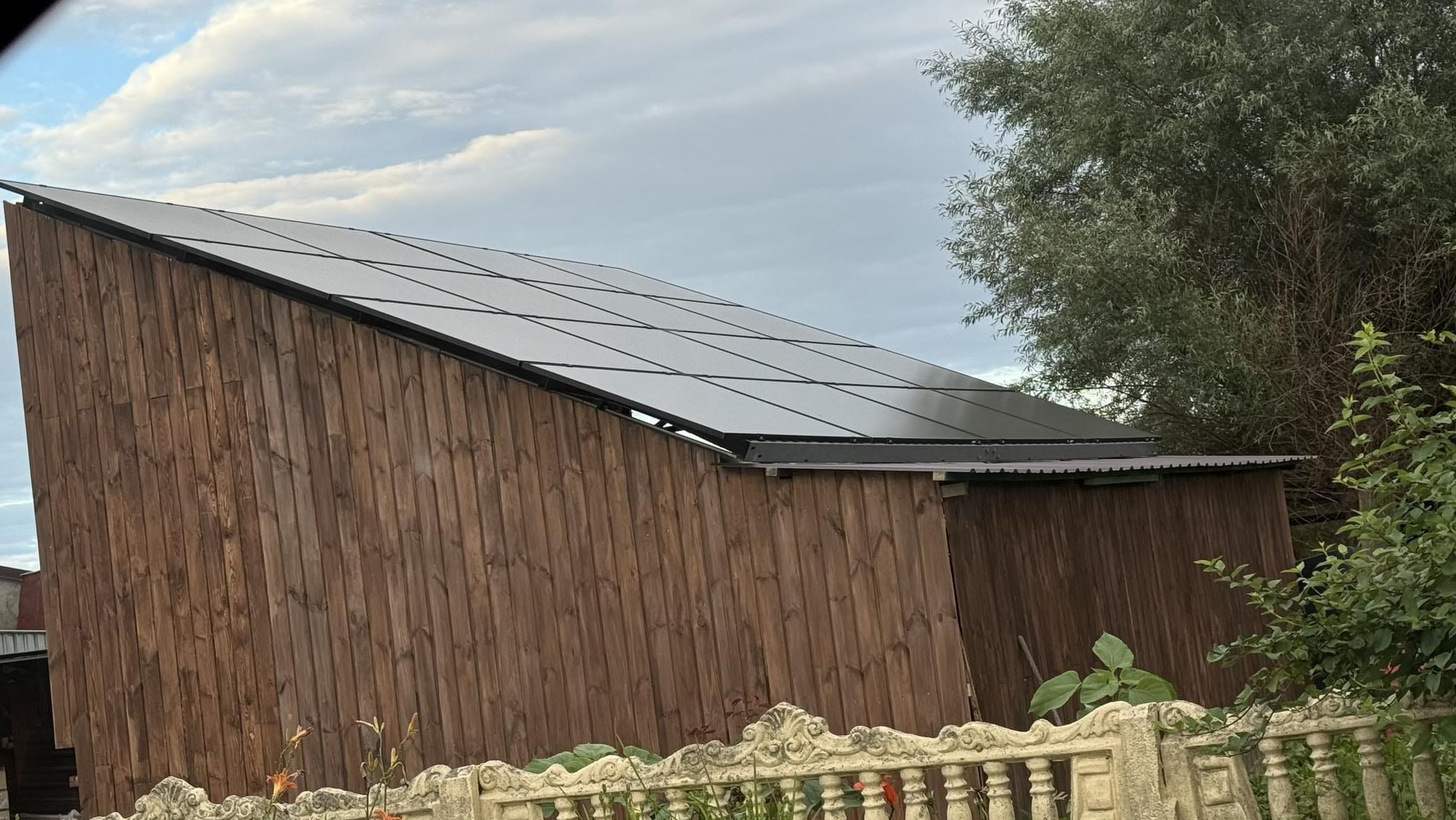 Panele słoneczne na skośnym dachu drewnianej konstrukcji, widok z dołu, fragment ozdobnego betonowego ogrodzenia w pierwszym planie.