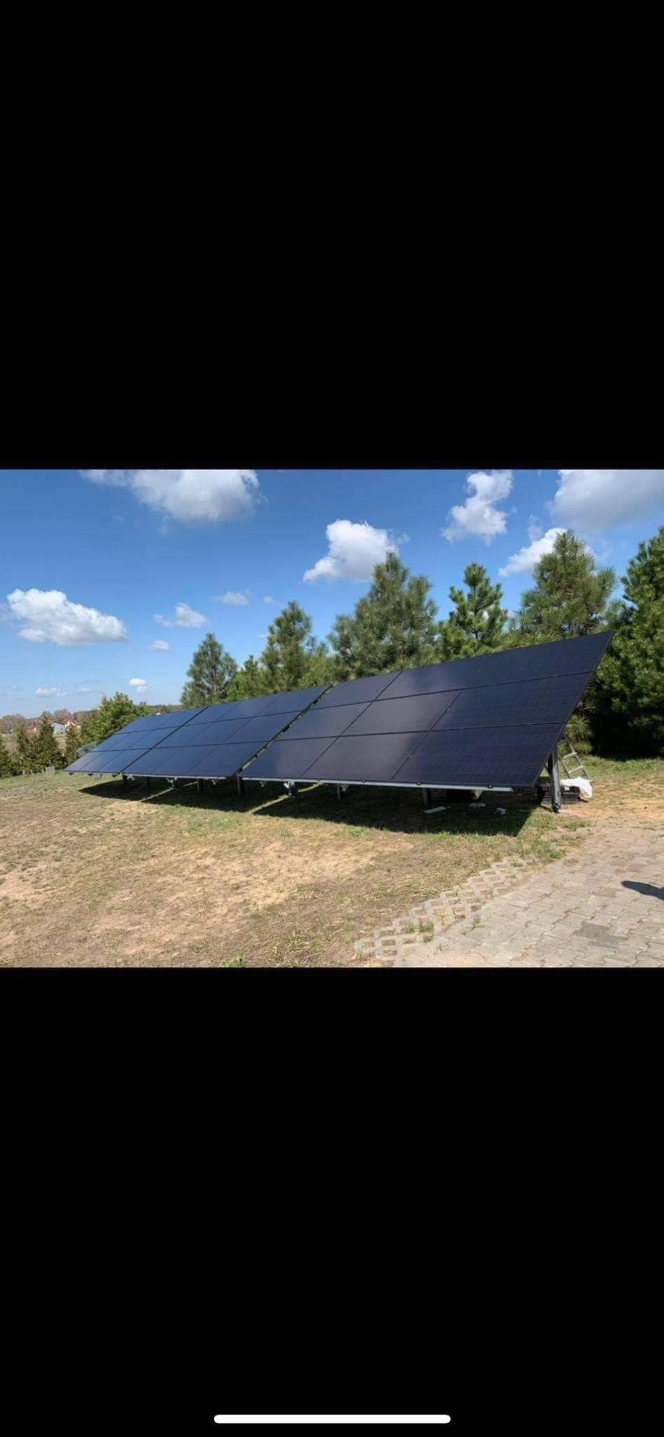 Rząd paneli słonecznych na trawiastym terenie z drzewami w tle, widoczna drabina i biała torba obok konstrukcji wsporczej.