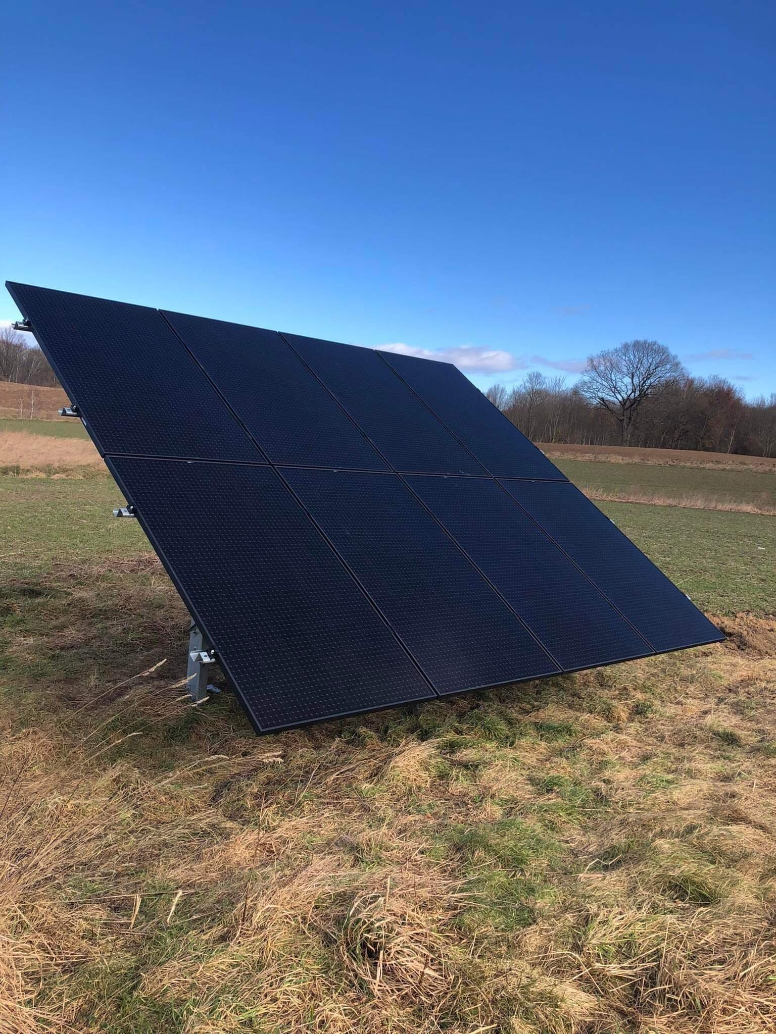 Czarny panel słoneczny ustawiony na metalowej konstrukcji na zewnątrz, na tle zielonego pola i błękitnego nieba z pojedynczym drzewem w oddali.