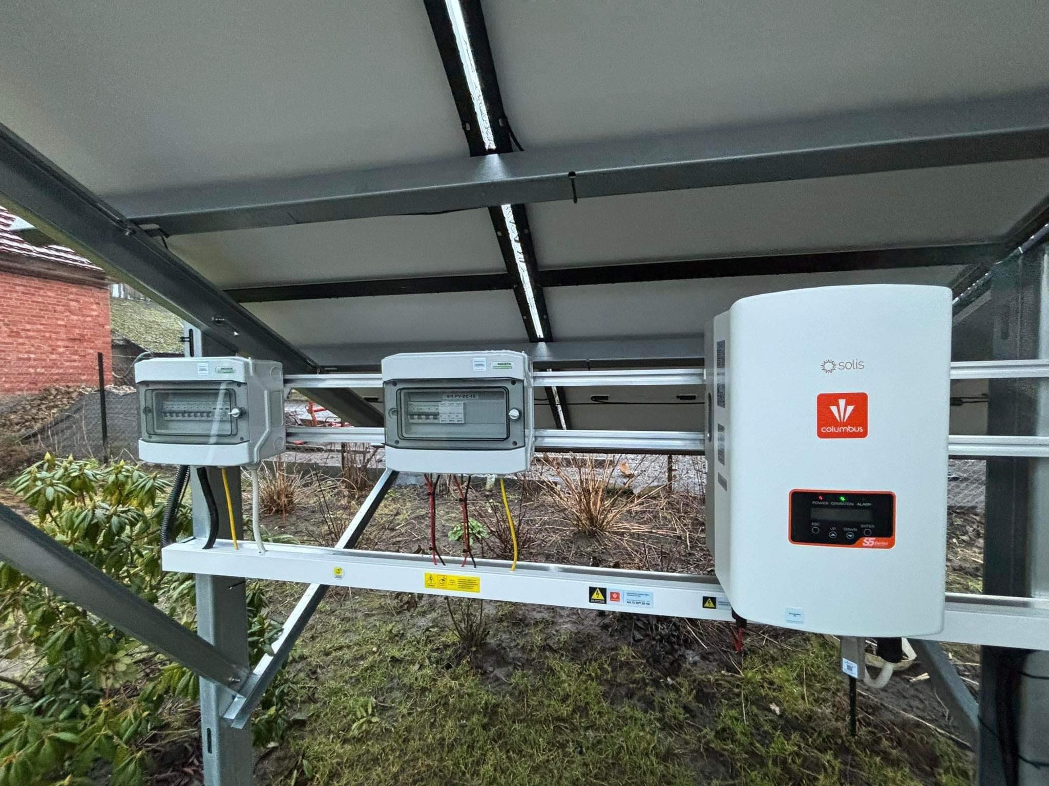 Instalacja fotowoltaiczna z widocznymi panelami, skrzynkami elektrycznymi i inwerterem Solis Columbus S5 na tle zieleni.
