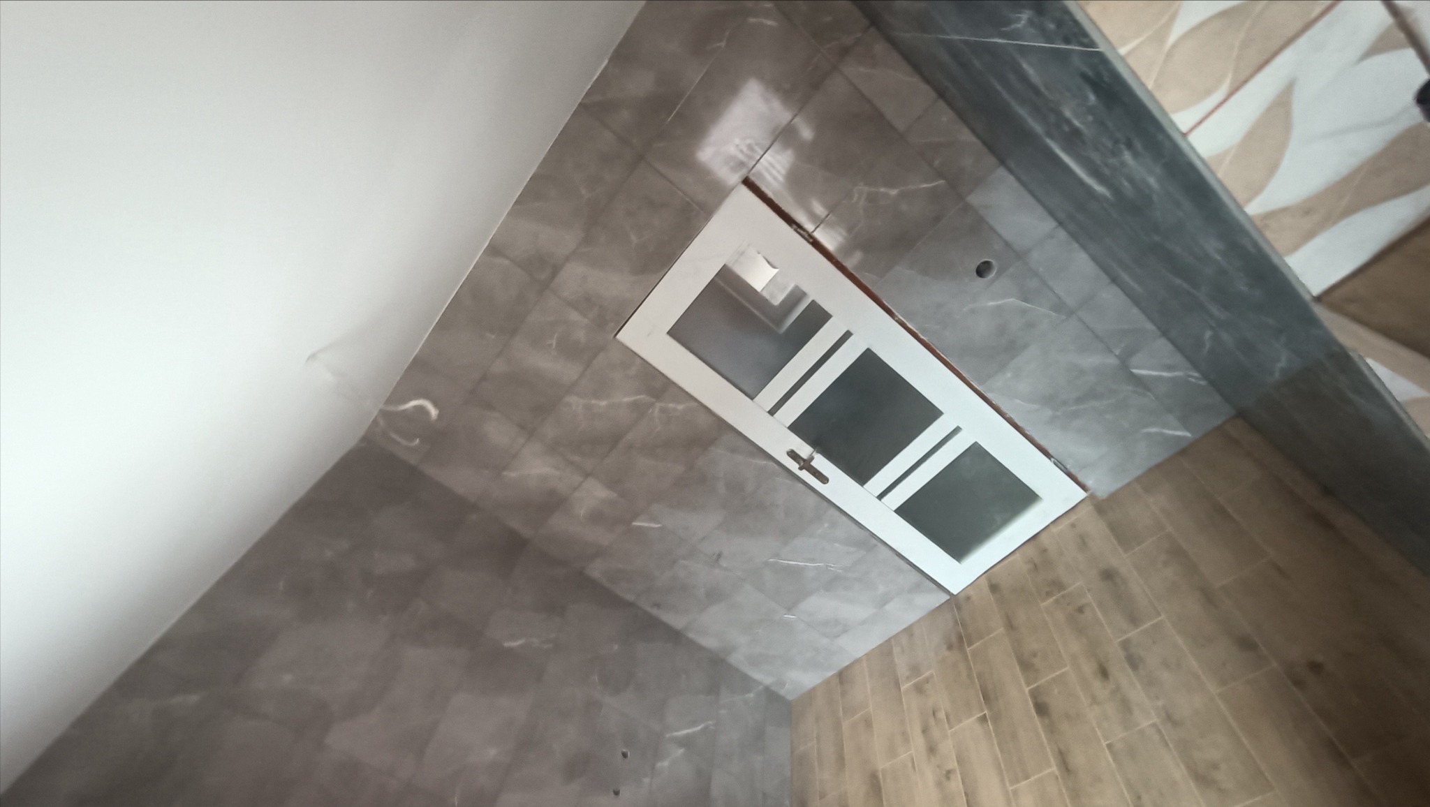 Ukończona łazienka z szarymi marmurkowymi płytkami na ścianach i drewnopodobnymi panelami na podłodze, widoczna biała rama drzwiowa z przeszkleniami.