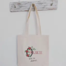Beżowa torba bawełniana z nadrukiem 'Polskie wie co dobre!' i motywem kwiatowym, zawieszona na wieszaku z postarzanego drewna.