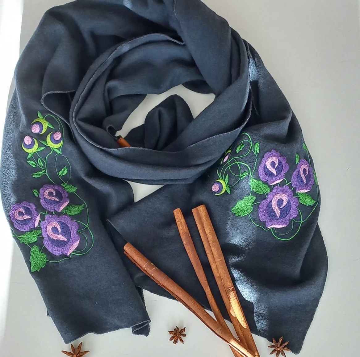 Grafitowy szal z haftowanymi fioletowymi różami i zielonymi pąkami, otoczony laskami cynamonu i anyżem gwiaździstym na białym tle.