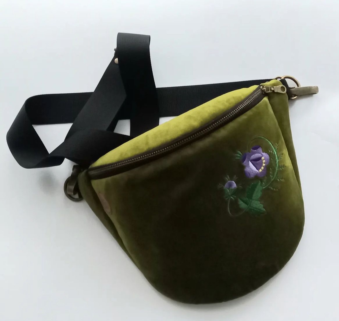 Zielona, aksamitna nerka z wyhaftowanym motywem fioletowego kwiatu i zielonych liści, leżąca na białym tle, z widocznym czarnym paskiem.
