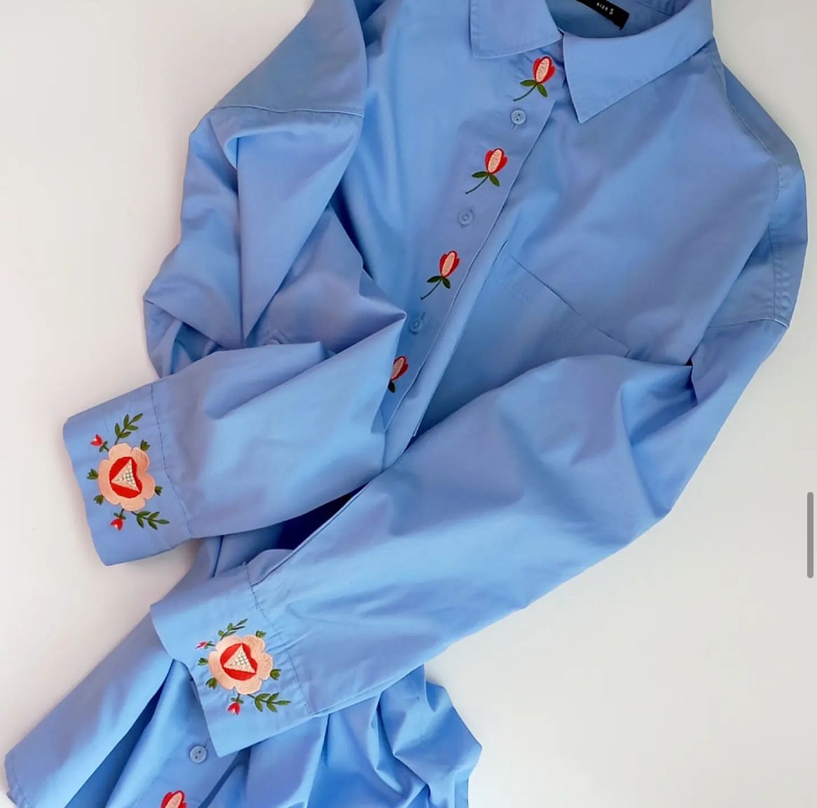 Jasnoniebieska koszula z długim rękawem, zdobiona haftowanymi motywami kwiatowymi wzdłuż zapięcia i na mankietach, leżąca na białym tle.