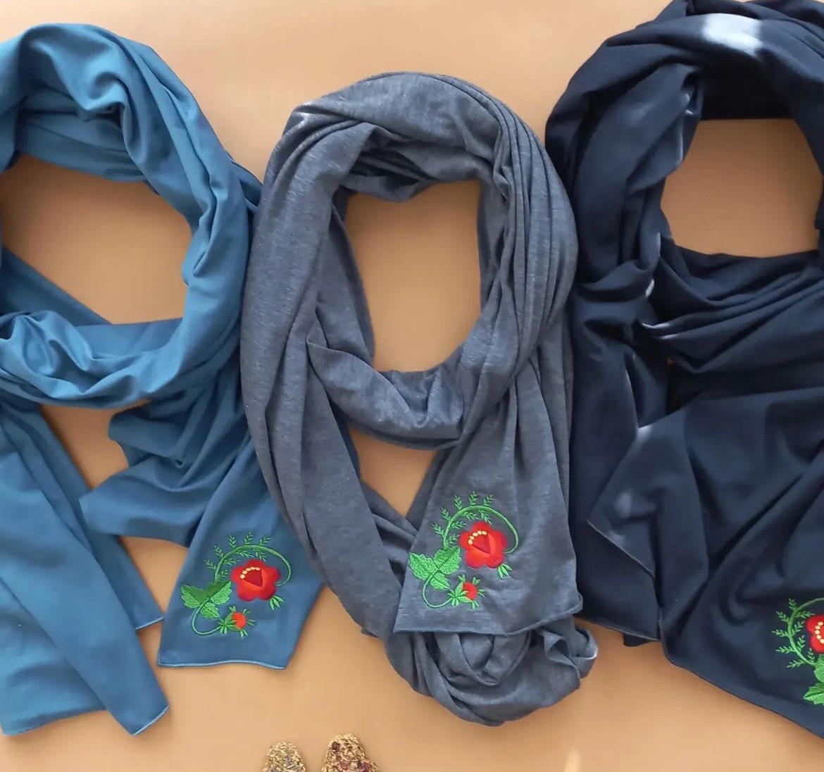 Trzy dzianinowe szale w odcieniach niebieskiego i szarości, każdy ozdobiony haftem przedstawiającym czerwoną różę z zielonymi listkami, ułożone na beżowym tle.
