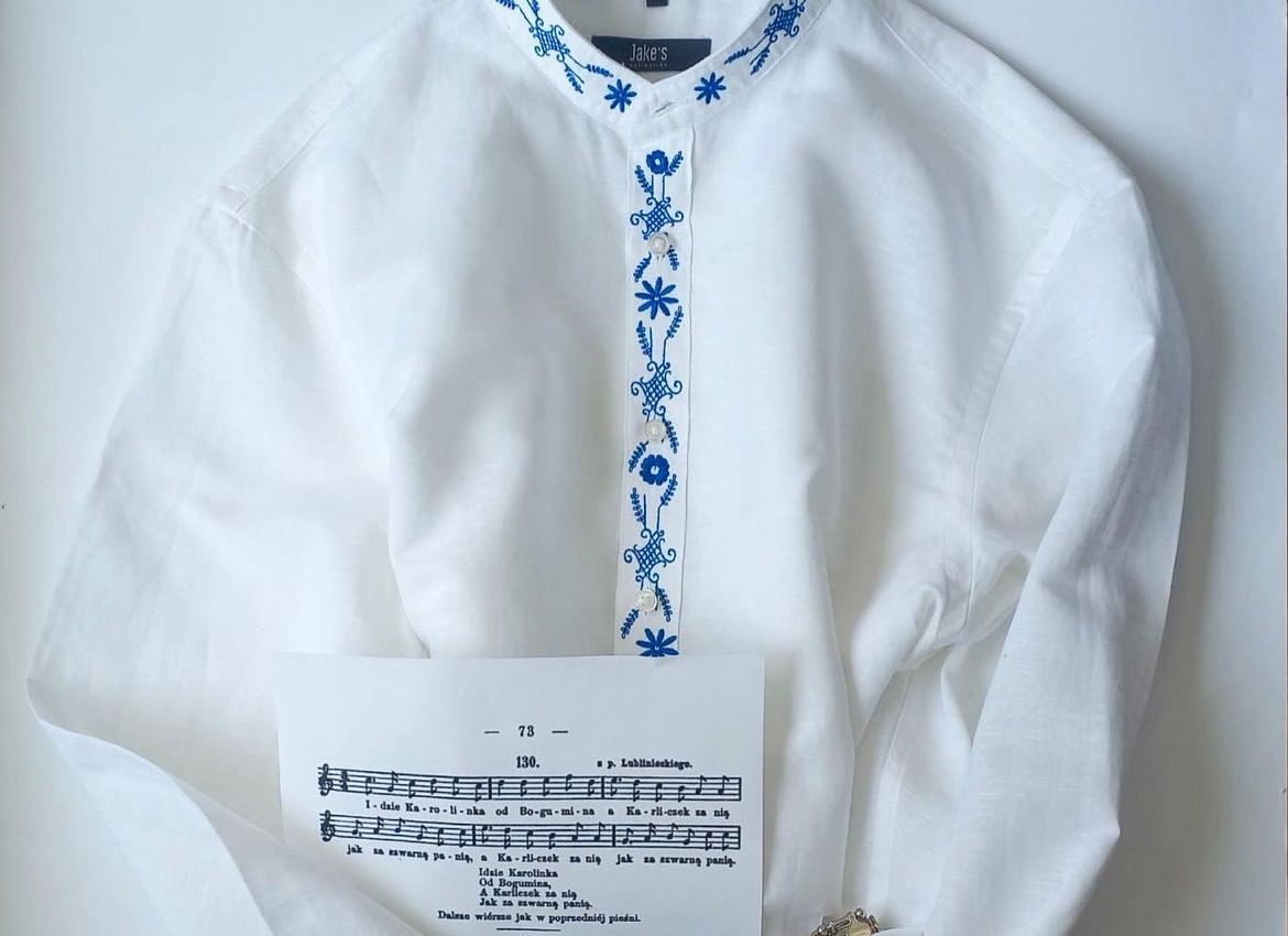 Biała koszula bez kołnierzyka z niebieskim haftem kwiatowym na listwie guzikowej i stójce, leżąca na białym tle obok kartki z nutami.