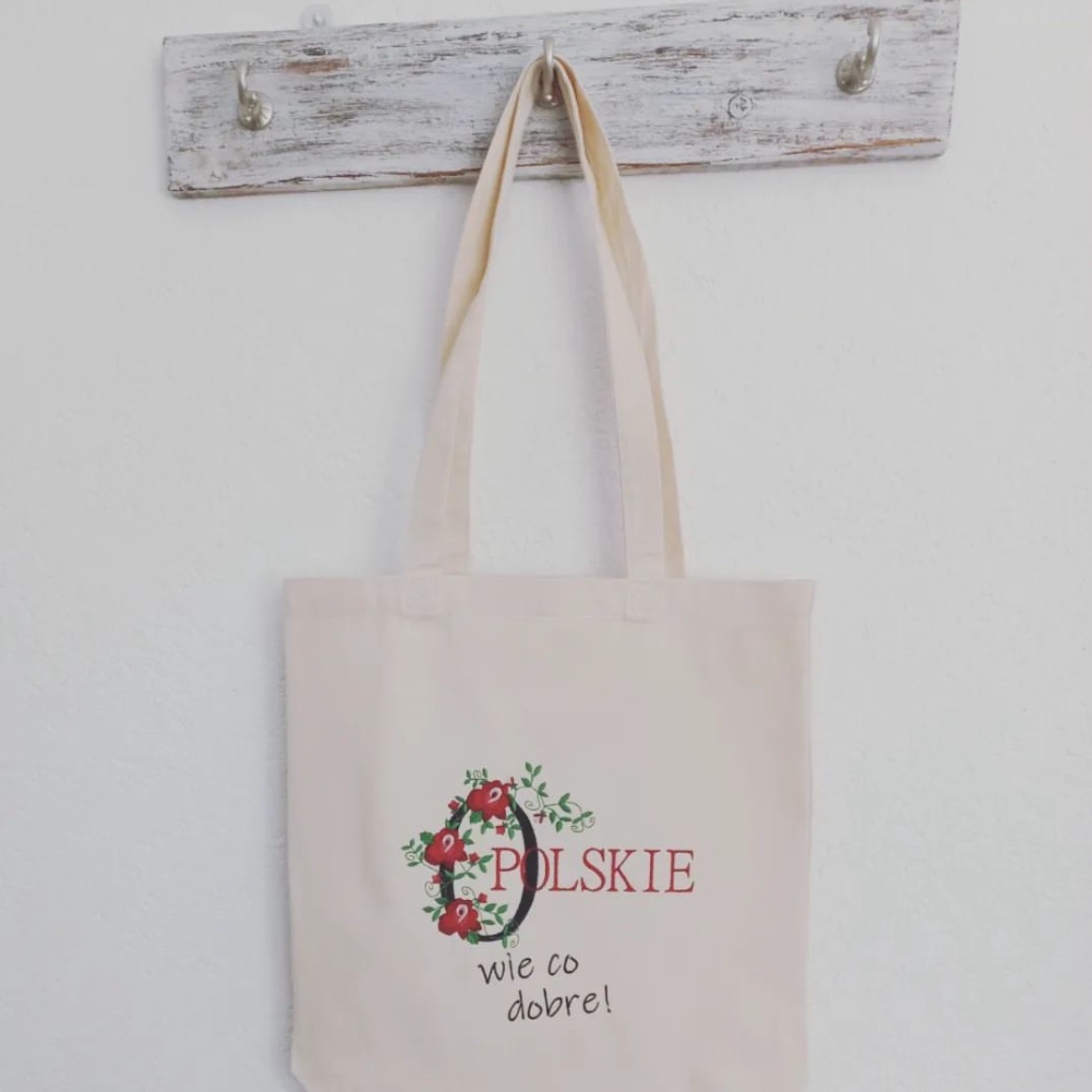 Beżowa torba bawełniana z nadrukiem 'Polskie wie co dobre!' i motywem kwiatowym, zawieszona na wieszaku z postarzanego drewna.