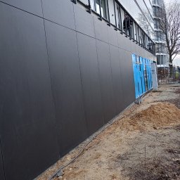 Firma Produkcyjno Handlowa Art-Dekor - Nowoczesna elewacja budynku biurowego z ciemnoszarych paneli, okna zabezpieczone niebieską folią ochronną, widoczny pracownik na balkonie i piasek budowlany u podstawy ściany.