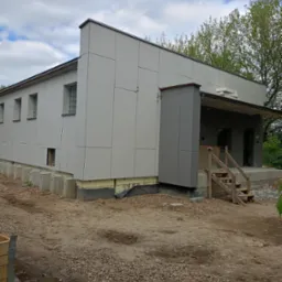 Budynek w trakcie montażu elewacji z paneli kompozytowych, widoczne betonowe podpory fundamentu, tymczasowe drewniane schody wejściowe i niezagospodarowany teren wokół budynku.