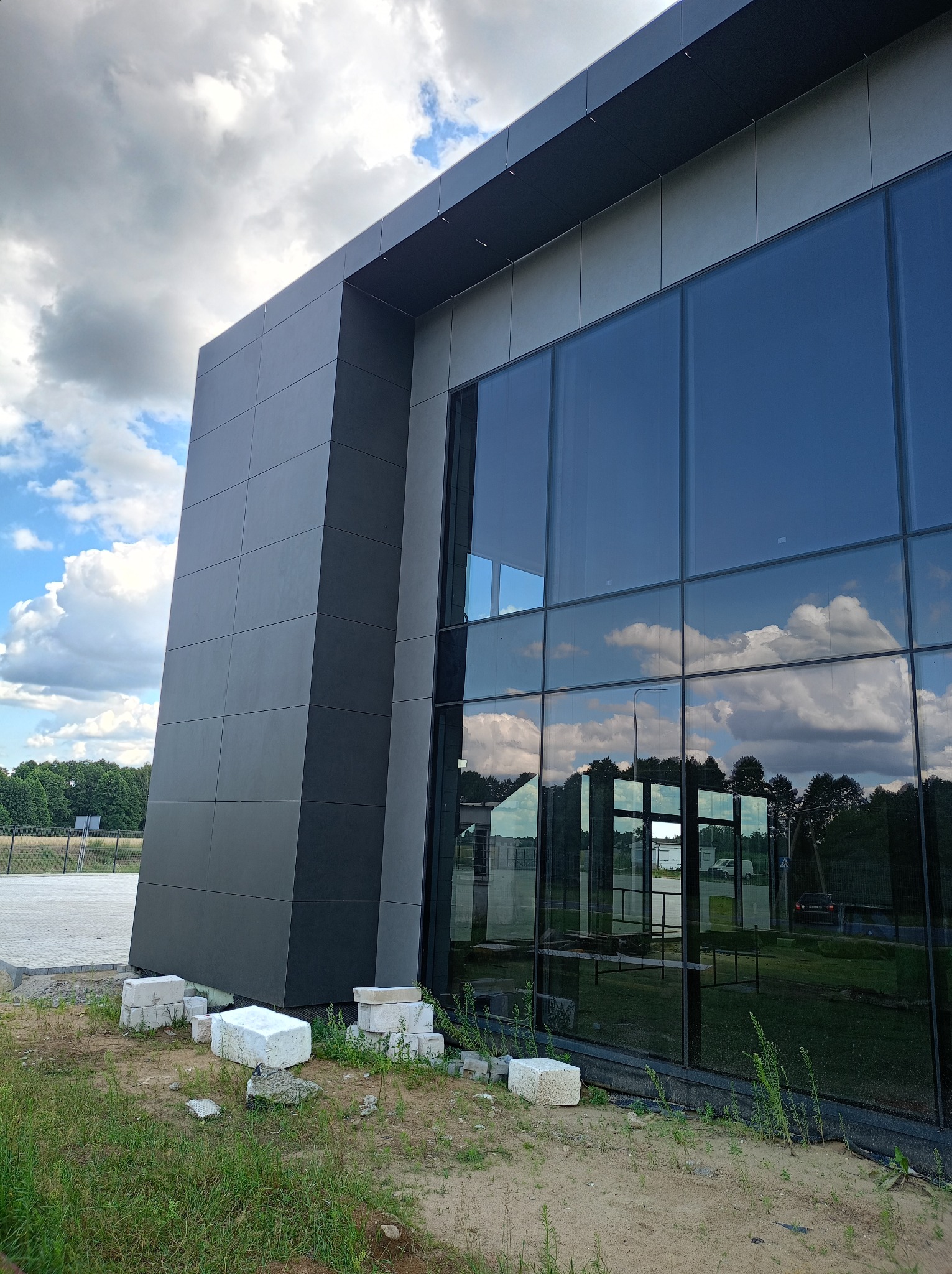 Nowoczesny budynek biurowy z elewacją z paneli w odcieniach szarości i dużymi oknami, w których odbija się błękitne niebo z chmurami. Na pierwszym planie, przed budynkiem, leżą białe bloki z betonu.