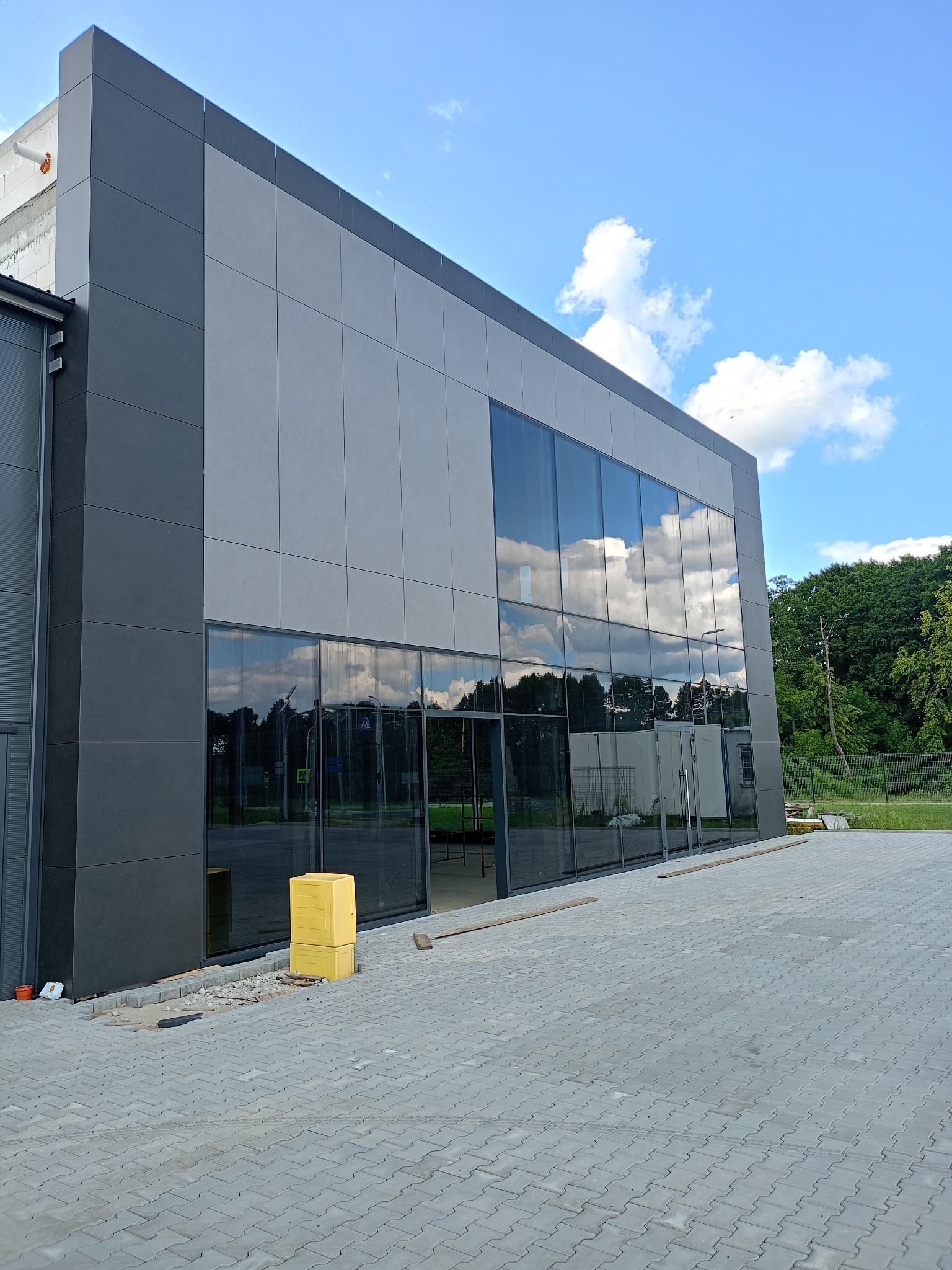 Nowoczesny budynek biurowy z szarą elewacją panelową i dużymi oknami odbijającymi niebo, widoczne niedokończone prace brukarskie przy wejściu.