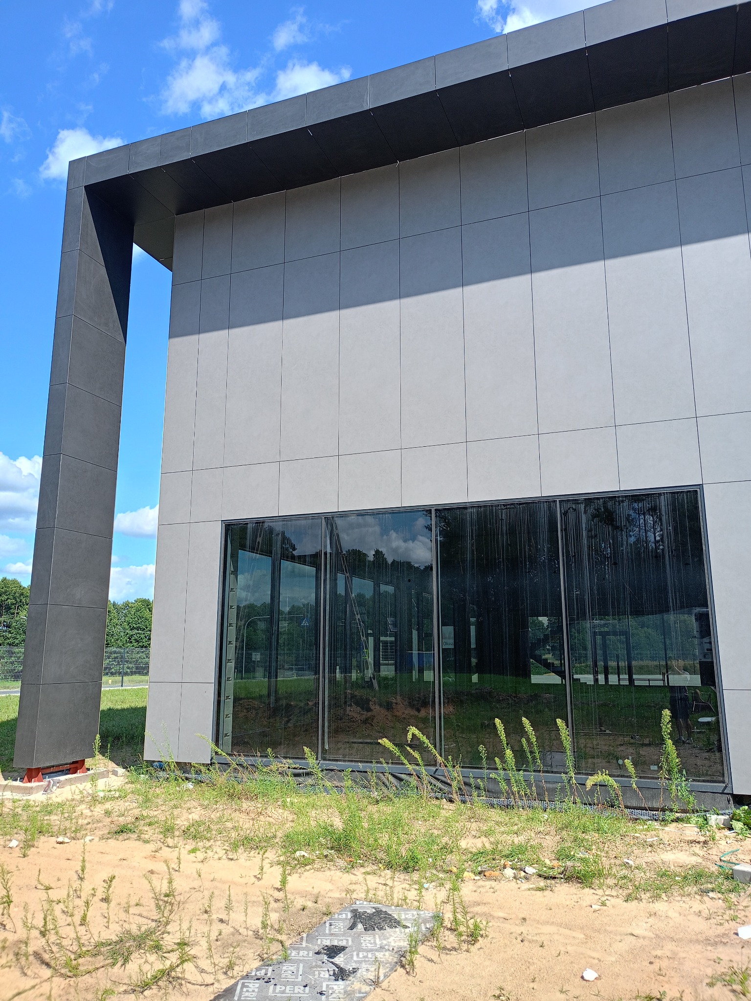 Nowoczesny budynek z elewacją z szarych płyt, widoczne duże okna z ciemnymi ramami i fragment terenu przed budynkiem z piaskiem i zieloną trawą.