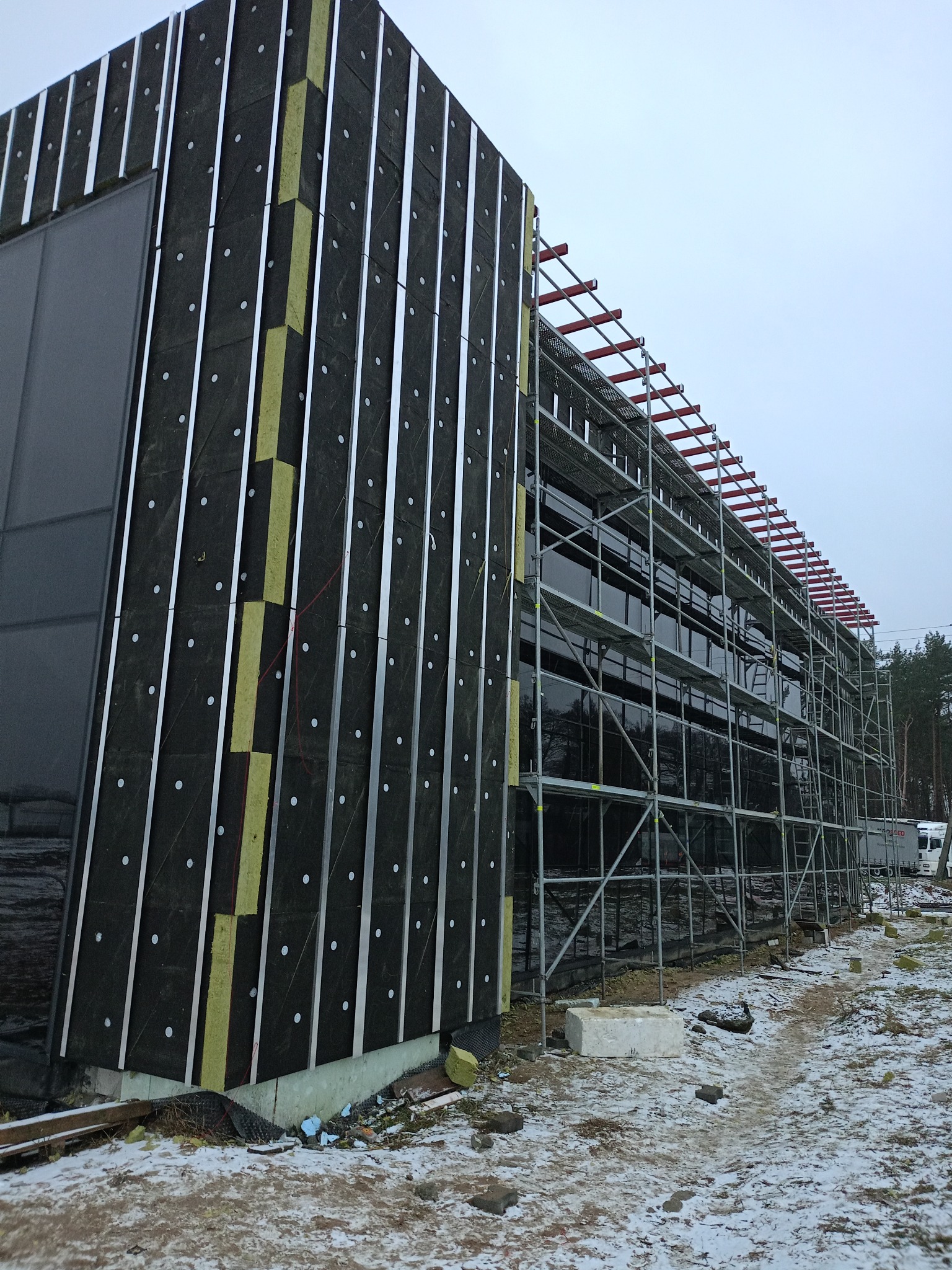 Budynek komercyjny w trakcie ocieplania: czarna membrana, wełna mineralna w pionowych pasach, metalowe profile, rusztowanie na całej ścianie, zimowa sceneria.
