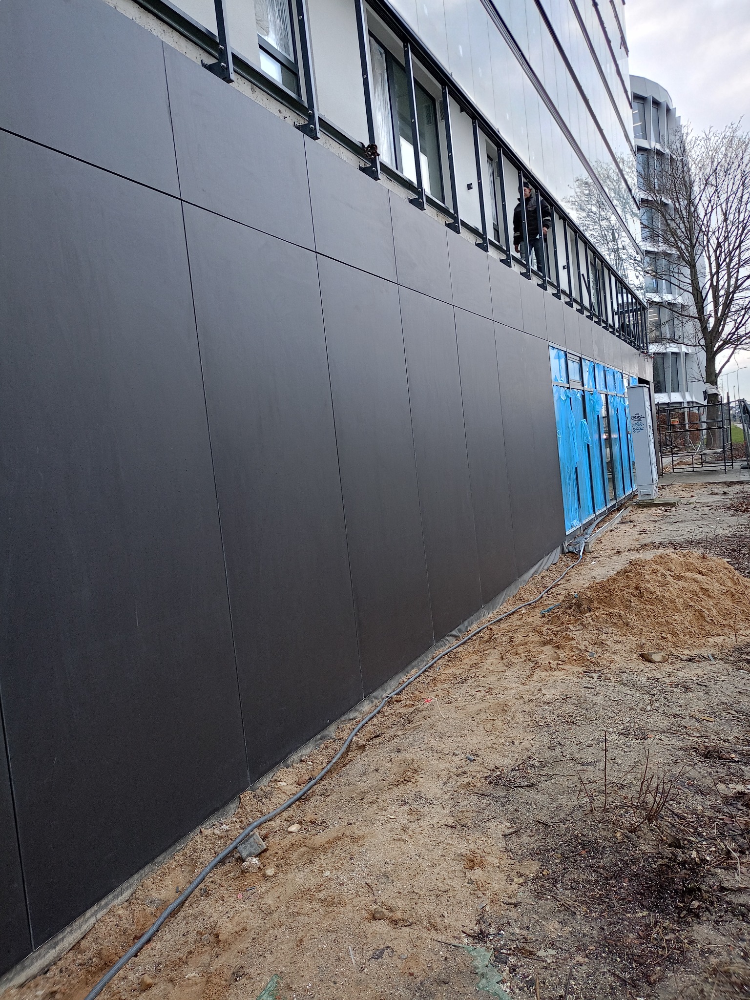 Nowoczesna elewacja budynku biurowego z ciemnoszarych paneli, okna zabezpieczone niebieską folią ochronną, widoczny pracownik na balkonie i piasek budowlany u podstawy ściany.