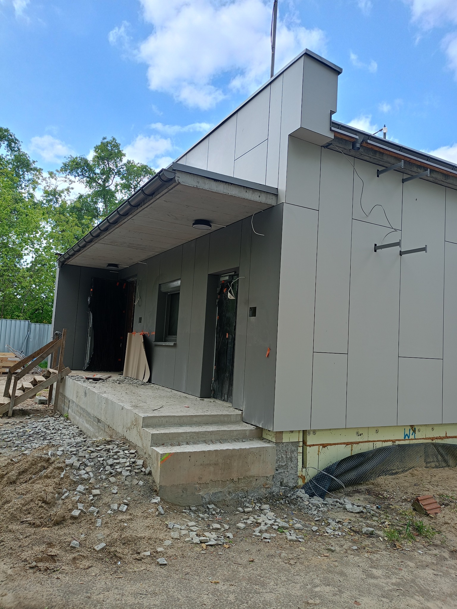 Budynek w trakcie montażu elewacji z szarych paneli, widoczne niedokończone wejście, schody z surowego betonu i tymczasowe drewniane schodki. W tle błękitne niebo z chmurami.