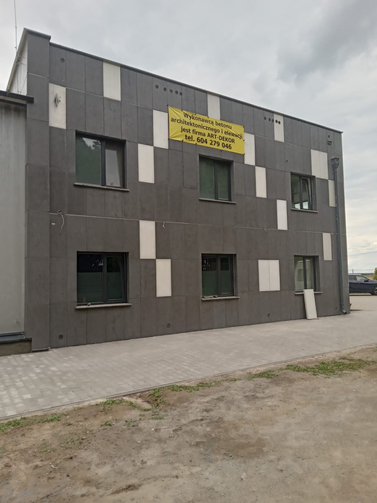 Budynek biurowy z szarą elewacją w geometryczny wzór z białymi akcentami, żółty baner reklamowy na górze elewacji, widoczne okna i chodnik z kostki brukowej.