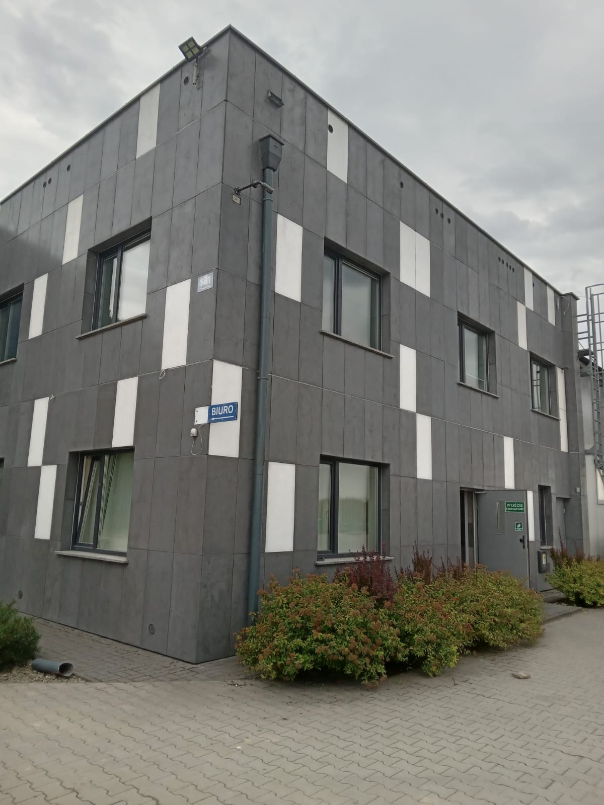 Nowoczesny budynek biurowy z szarą elewacją w białe pasy, widoczny znak 'Biuro' i krzewy przed wejściem.